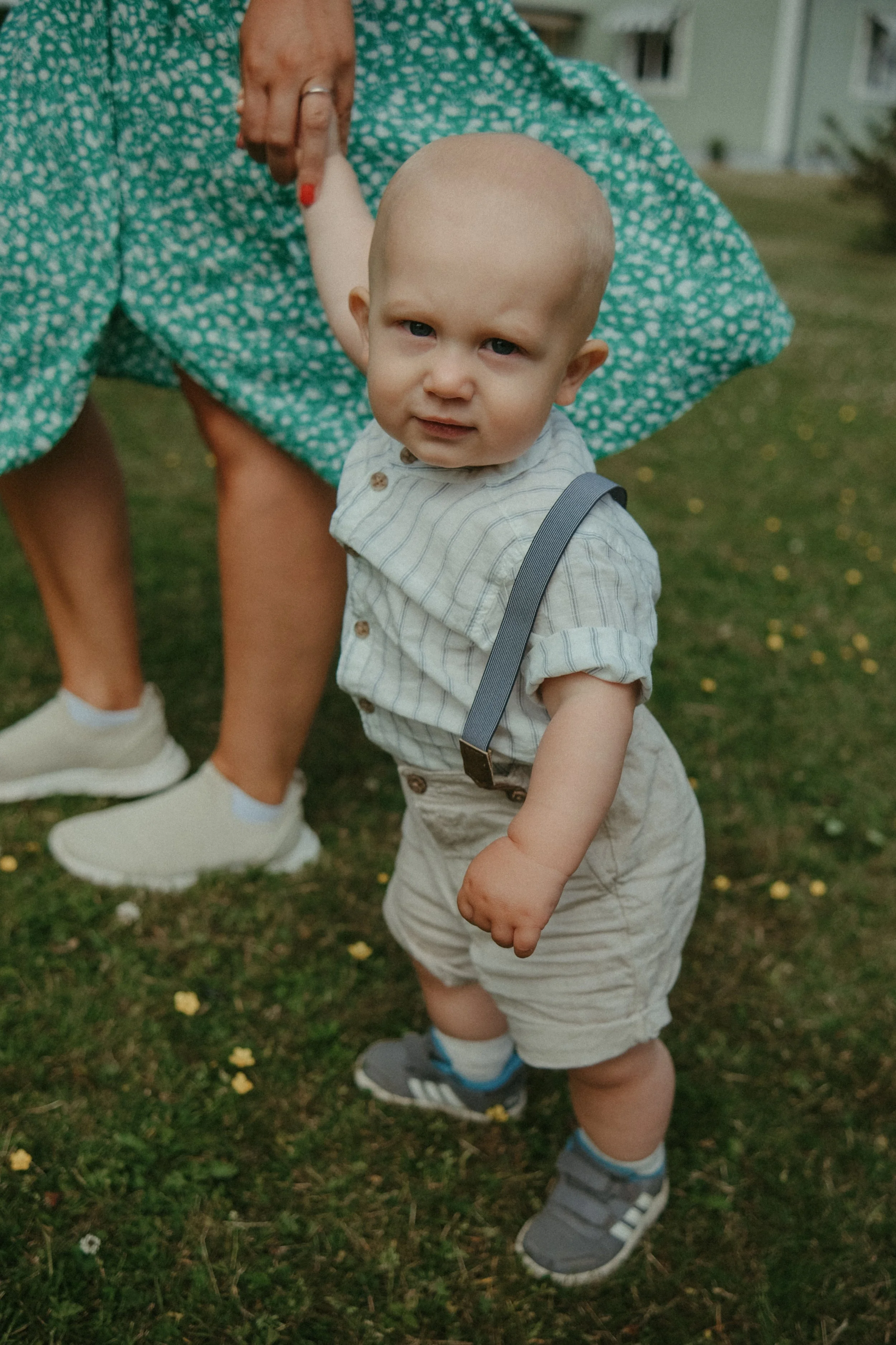 En ung pojke med blont hår, blå ögon och kortärmad skjorta, står på gräset. En kvinna, hans mamma, håller honom i handen. Hela scenen utspelar sig utomhus i en trädgård.