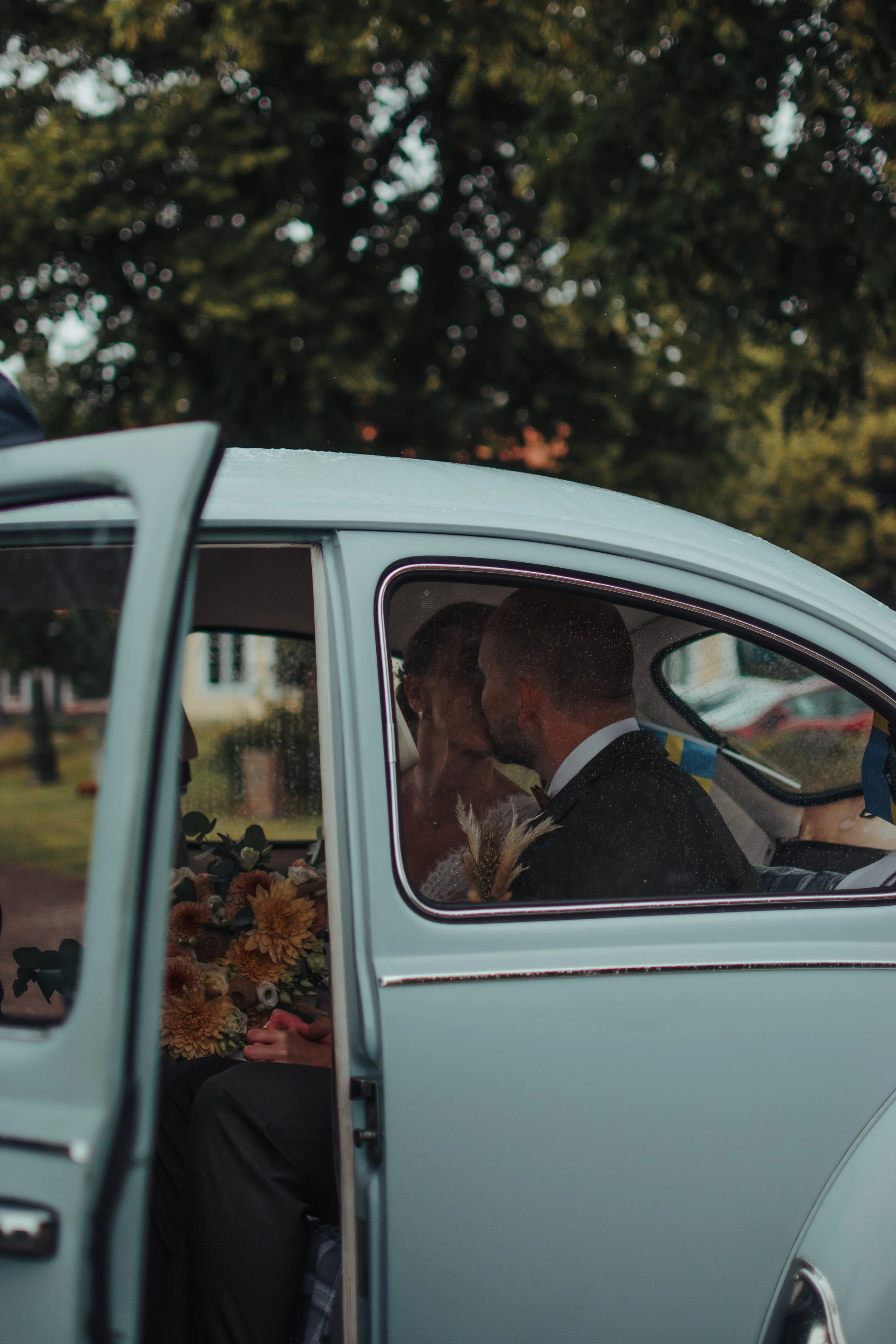 Två personer sitter i en vit bil med bakluckan öppnad, omgivna av blommor och träd i bakgrunden.