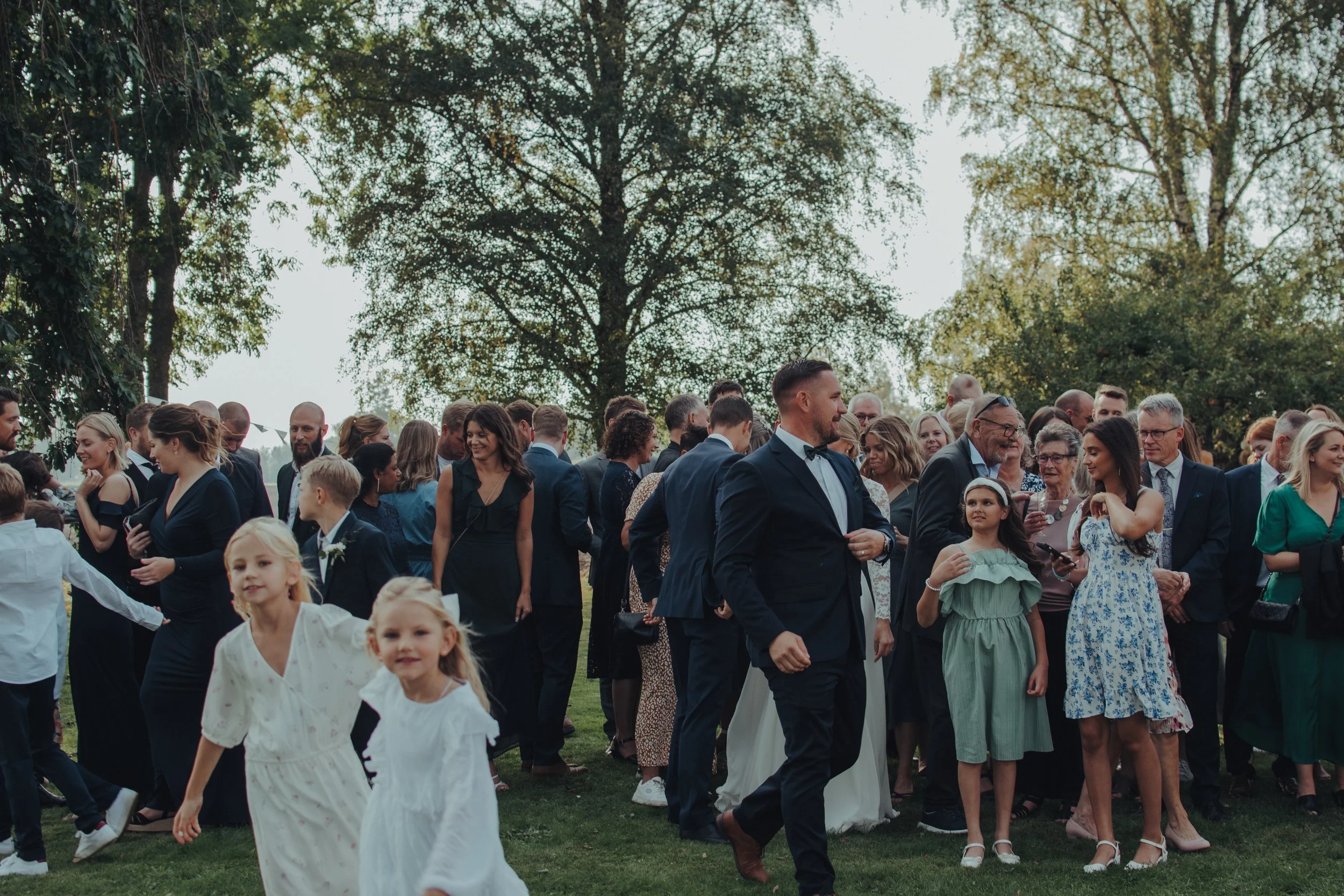 En stor grupp människor dansar och umgås utomhus under en dag med klart väder, omgiven av gröna träd.