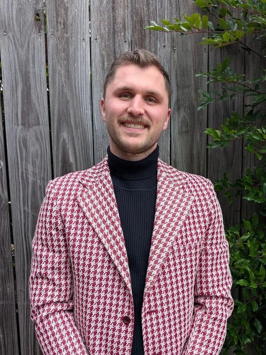 A man with short light brown hair and a beard, smiling, wearing a black turtleneck and a red and white houndstooth blazer, standing outdoors in front of a wooden fence and green foliage.
