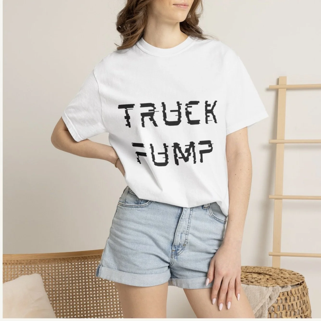 A woman wearing a white T-shirt with the words "TRUCK FUMP" written in a distorted, glitchy font and light denim shorts, standing indoors near a woven chair and a wooden ladder.