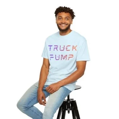 A smiling man with curly hair and a beard sitting on a black stool, wearing a light blue T-shirt with the words 'TRUCK PUMP' written in colorful letters.