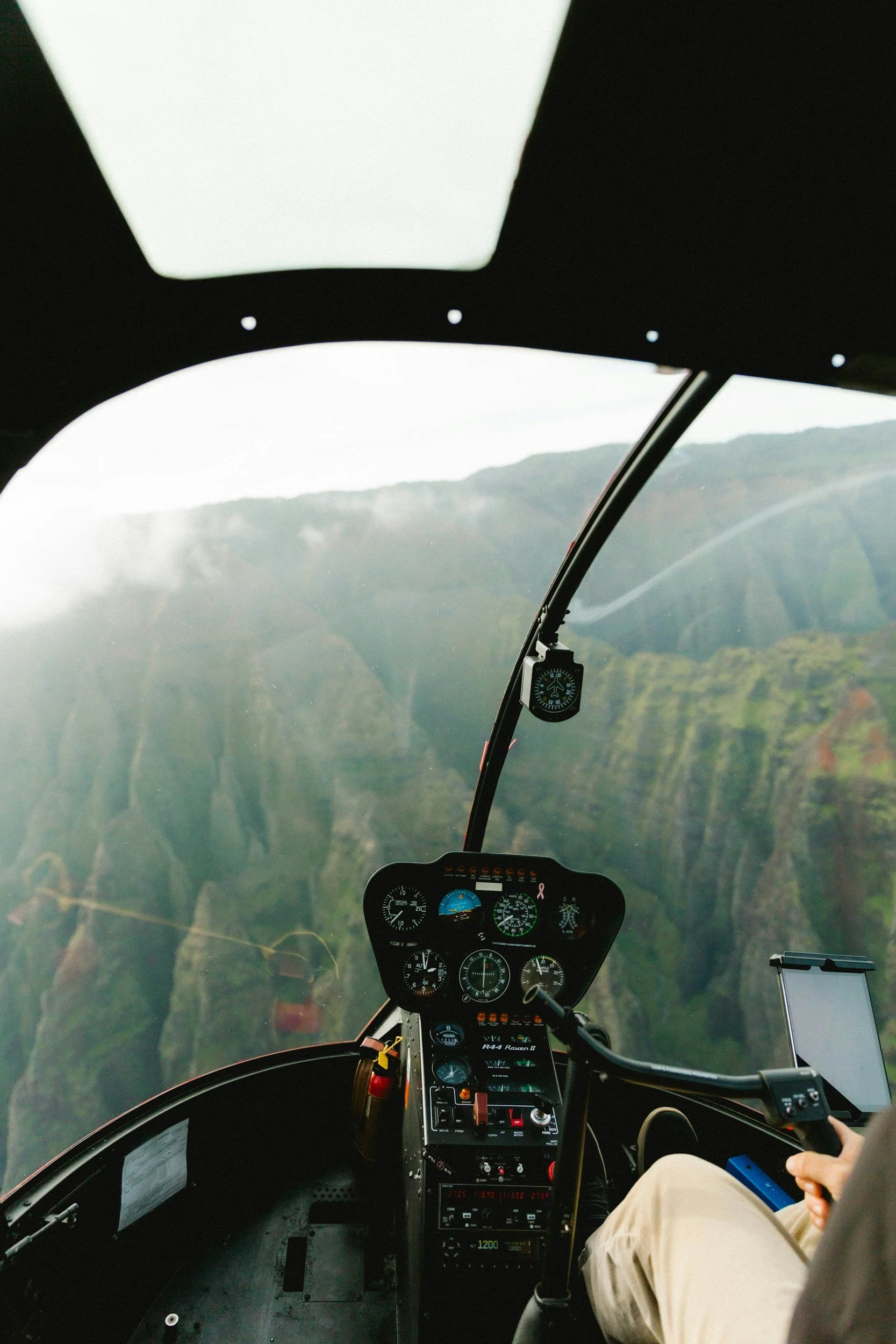 Interior view of a helicopter cockpit with various instruments and gauges, with mountains and cloudy sky visible through the windshield.