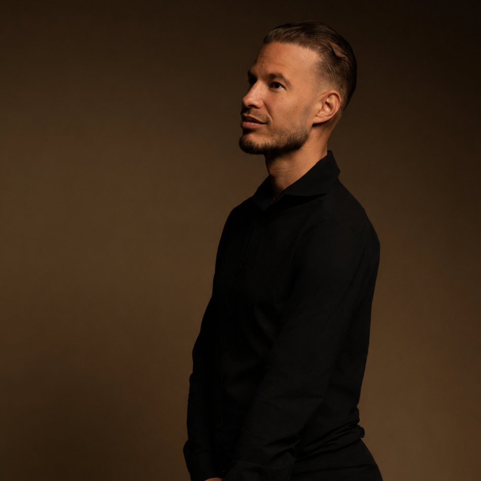 Portret van een man met een gekapte haarstijl en een baard, gekleed in een zwarte blouse, staat tegen een effen, donkere achtergrond.