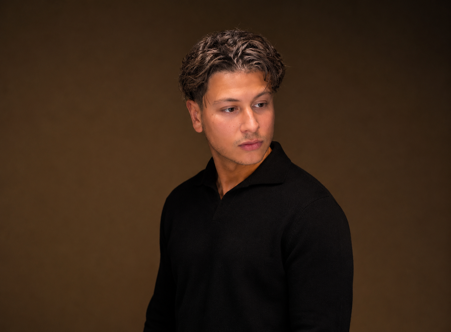 Portret van een jonge man met kort, golvend haar, gekleed in een zwart shirt, kijkend naar links tegen een effen bruine achtergrond.