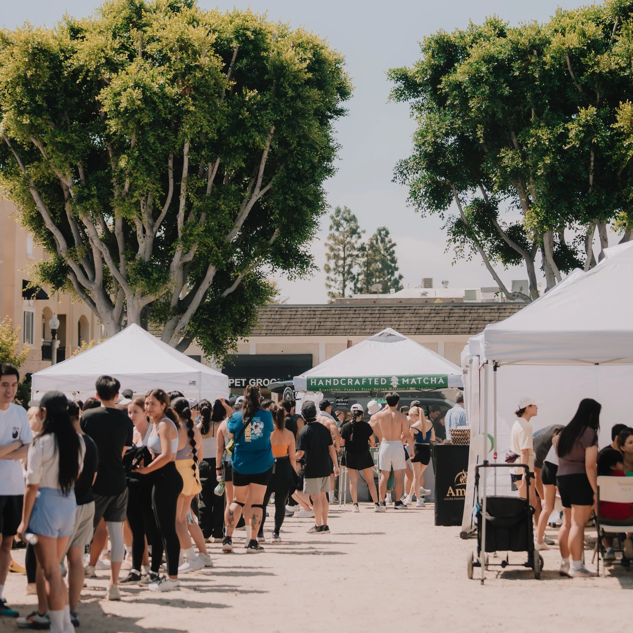 Crowd of people walking outdoors at an event, with white tents and green trees in the background.