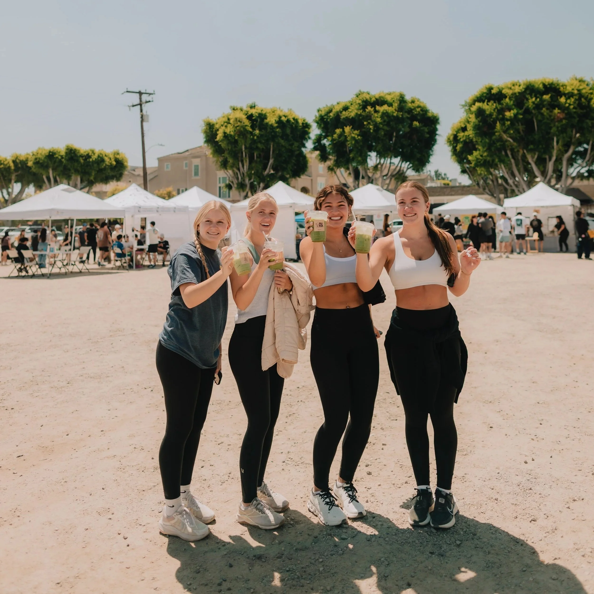 Four young women standing outdoors at a community event, smiling and holding large cups of green smoothies. They are dressed in casual athletic wear, with tents and crowd in the background, under clear skies and leafy trees.