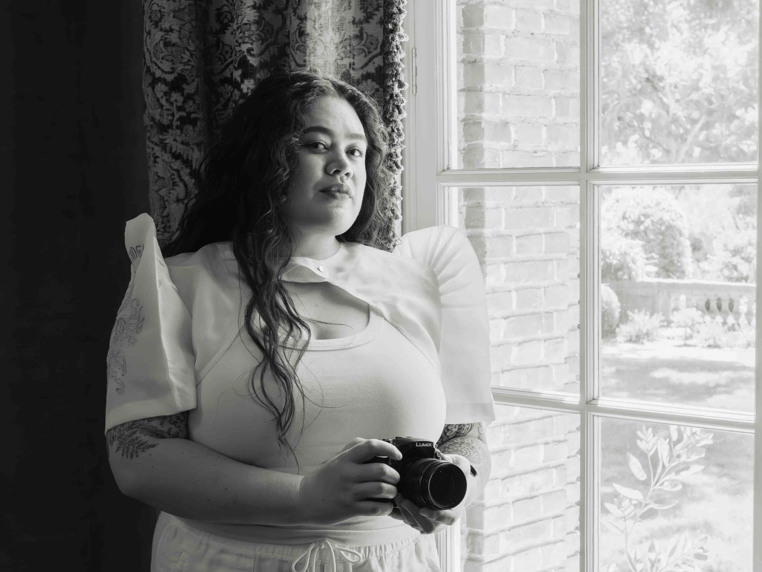 A woman with curly hair resting on a windowsill, holding a camera, with a brick wall visible outside the window.