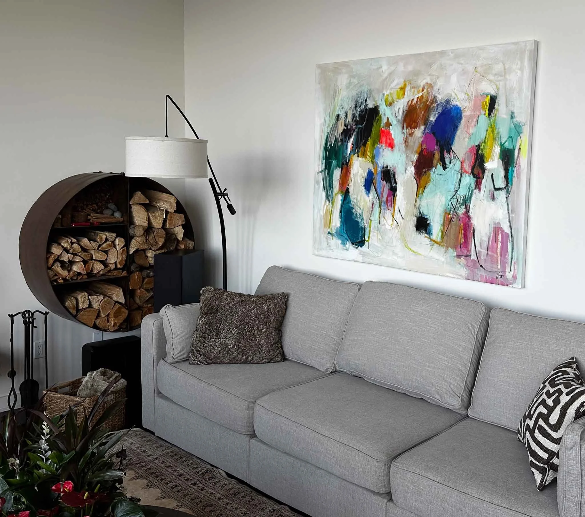 A living room with a gray couch featuring a dark textured pillow, a large colorful abstract painting on the wall, a black floor lamp, a wood log firewood holder, and a potted plant with pink flowers.