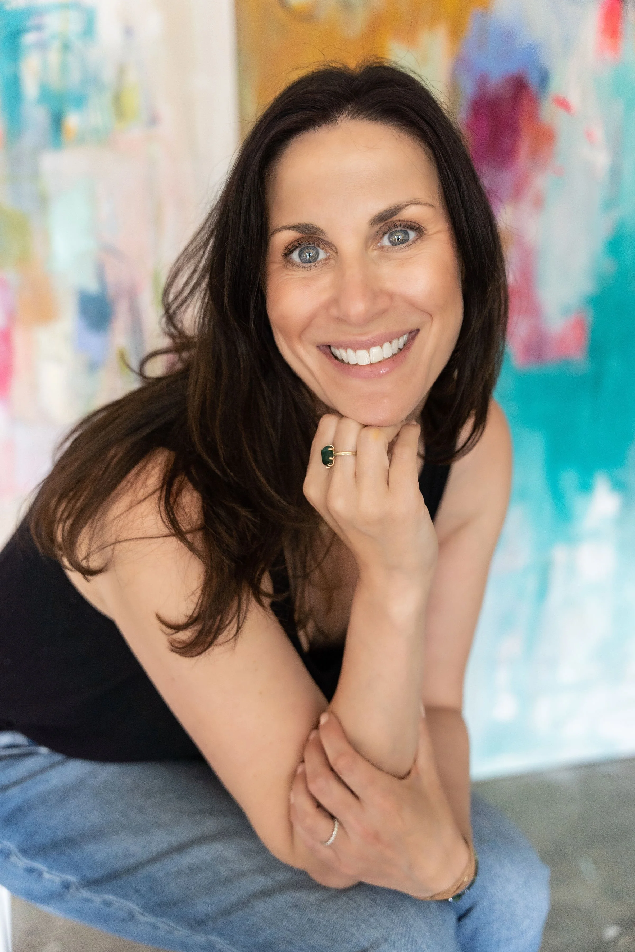 Close-up of a smiling woman with long dark hair and blue eyes, wearing a black top and gray jeans, sitting in front of a colorful abstract background.