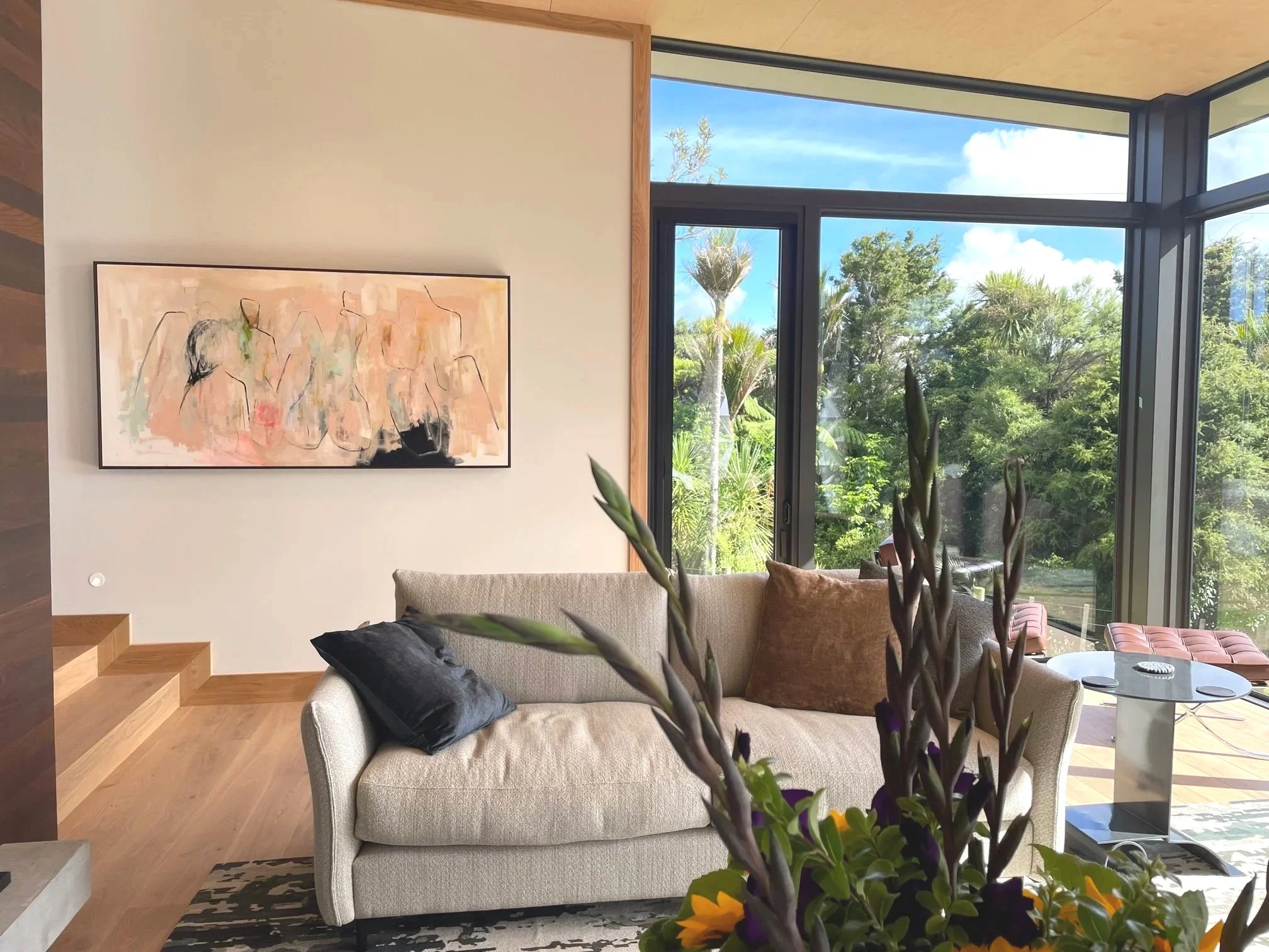 Modern living room with large windows showing green trees and blue sky outside. Beige sofa with black and brown pillows, abstract wall art, and indoor plants in the foreground. Wooden stairs to the left.
