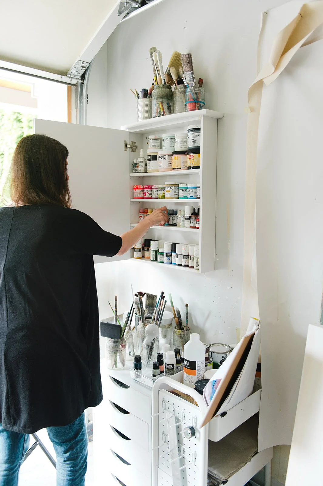 A person organizing bottles of paint and art supplies in a white cabinet in an art studio.