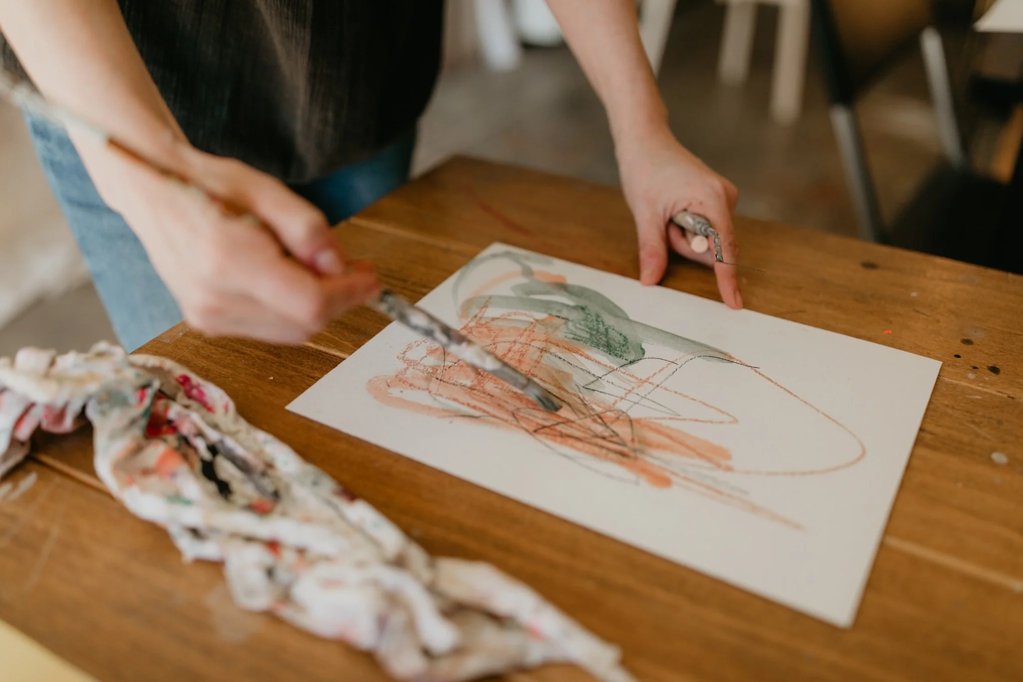 A person holding a paintbrush and painting on a piece of paper on a wooden table. There is a crumpled paper towel with paint on it nearby.