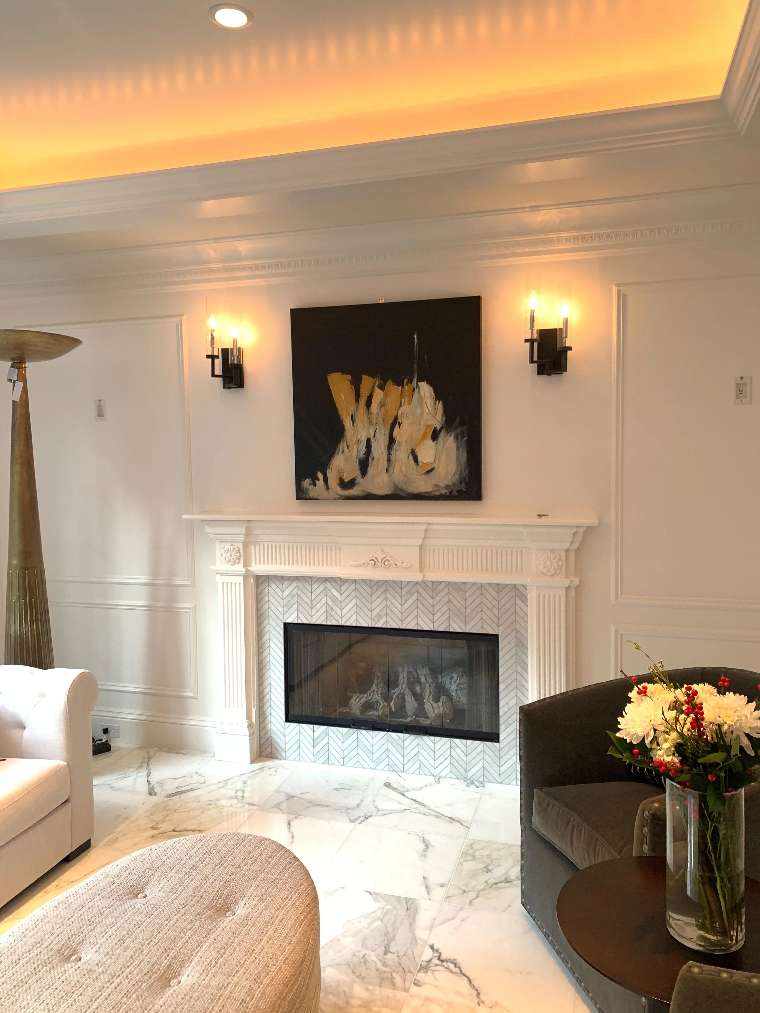 Living room with white walls, marble floor, a fireplace with a herringbone-patterned tile surround, wall sconce lights, a framed artwork above the fireplace, and a flower arrangement on a small table.