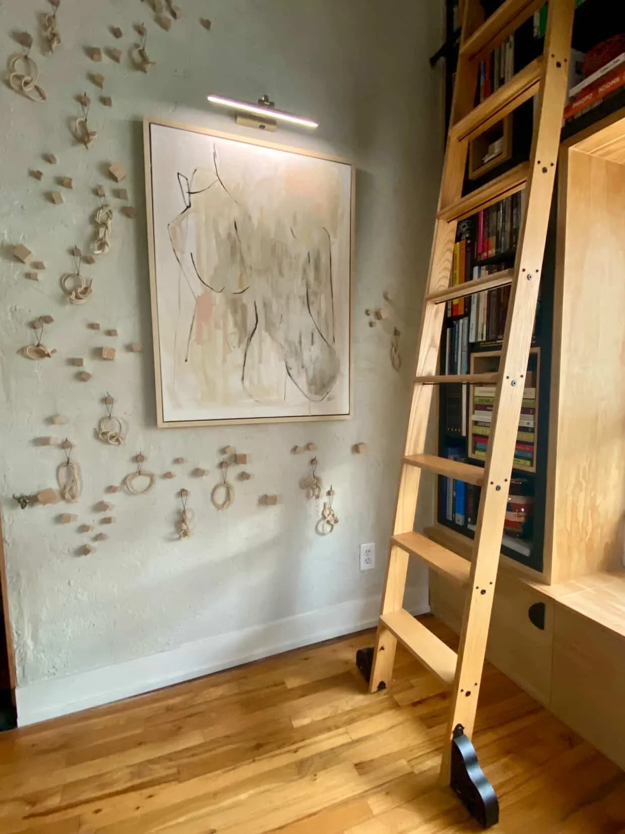 Interior view of a room with a light-colored wall, a wooden bookshelf with books, a ladder leaning against the bookshelf, a large abstract art piece on the wall, and decorative wooden rings hanging on the wall.
