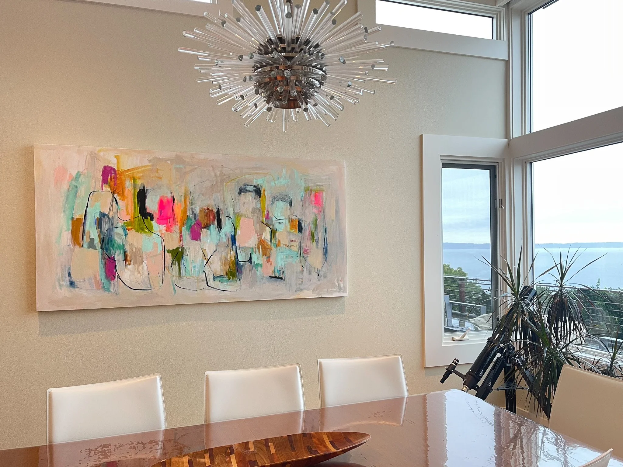 A modern dining room with a multicolored abstract painting on a cream wall, a wooden dining table with white chairs, large windows with white trim, and a starburst ceiling light fixture.