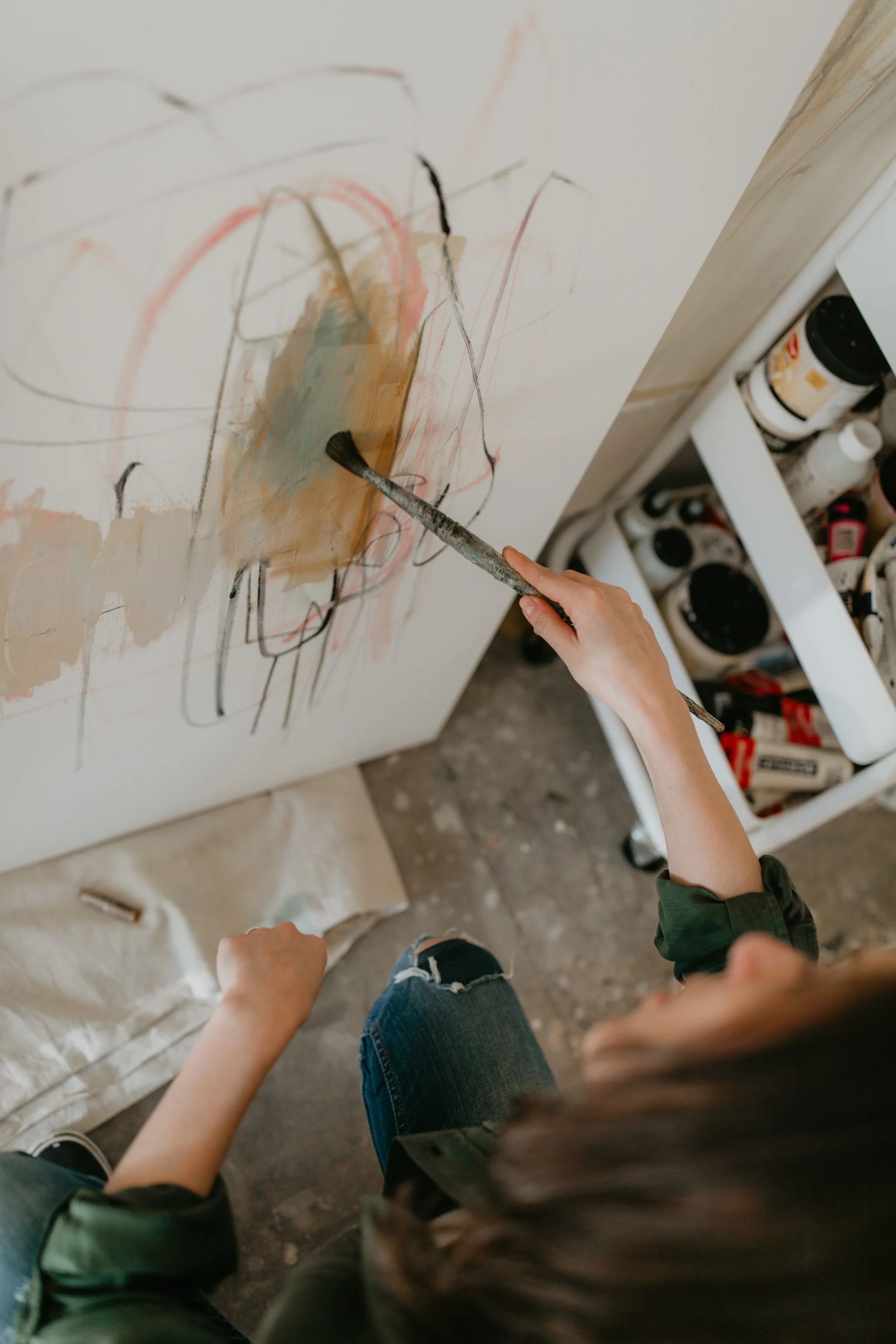 Person pointing at a partially painted abstract artwork on a large white canvas. The scene is in an art studio with paint bottles and supplies visible.