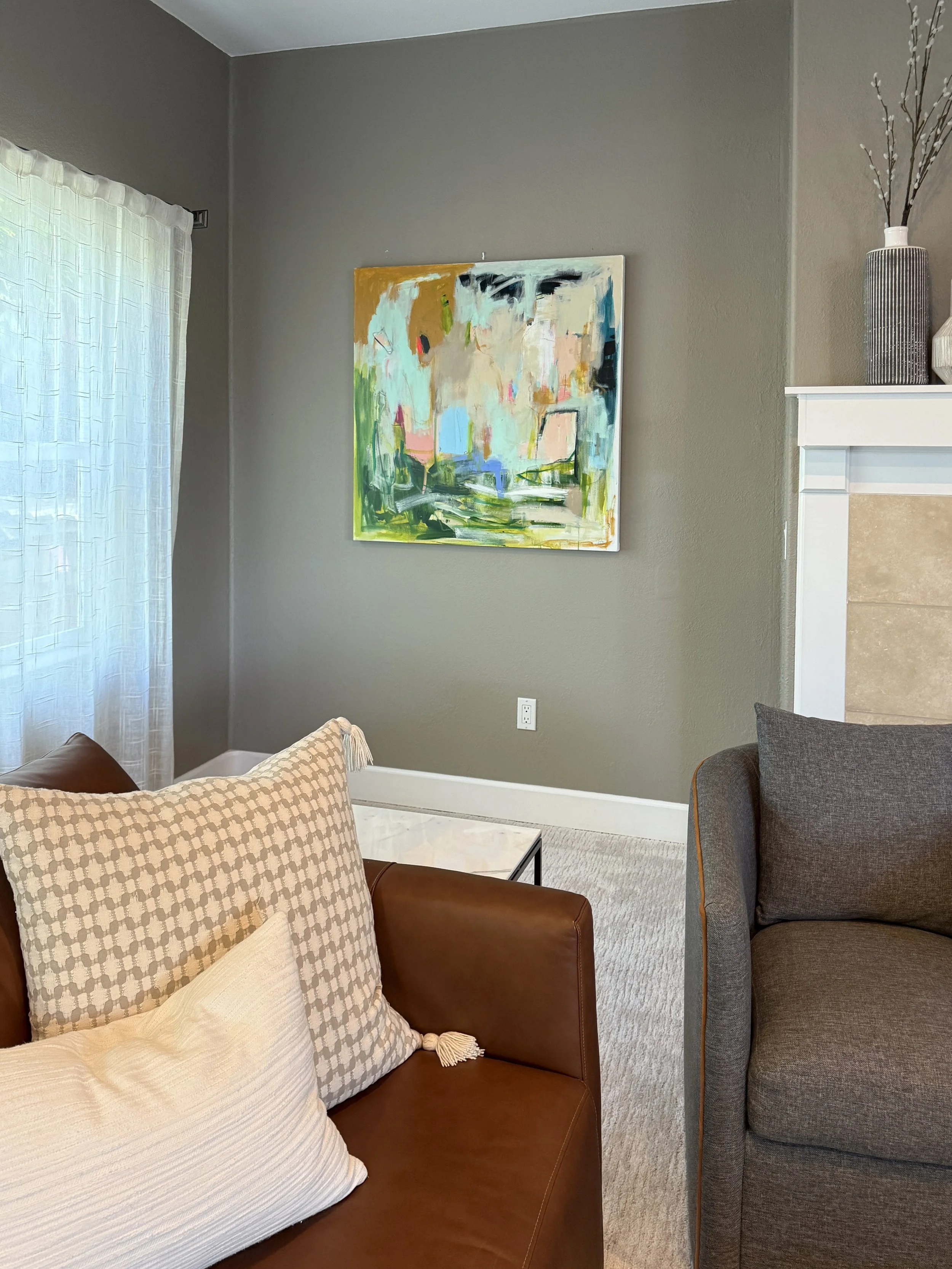 A corner of a living room with a gray wall, abstract art painting, a window with white curtains, a brown leather couch with pillows, and a gray armchair.