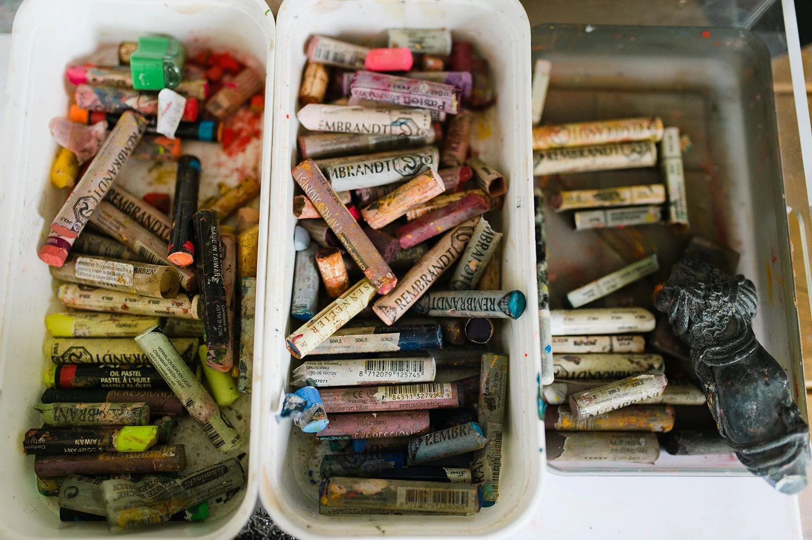 Containers filled with used oil pastels in various broken and worn condition, with colors including pink, yellow, green, black, and blue, some with visible labels and barcodes.