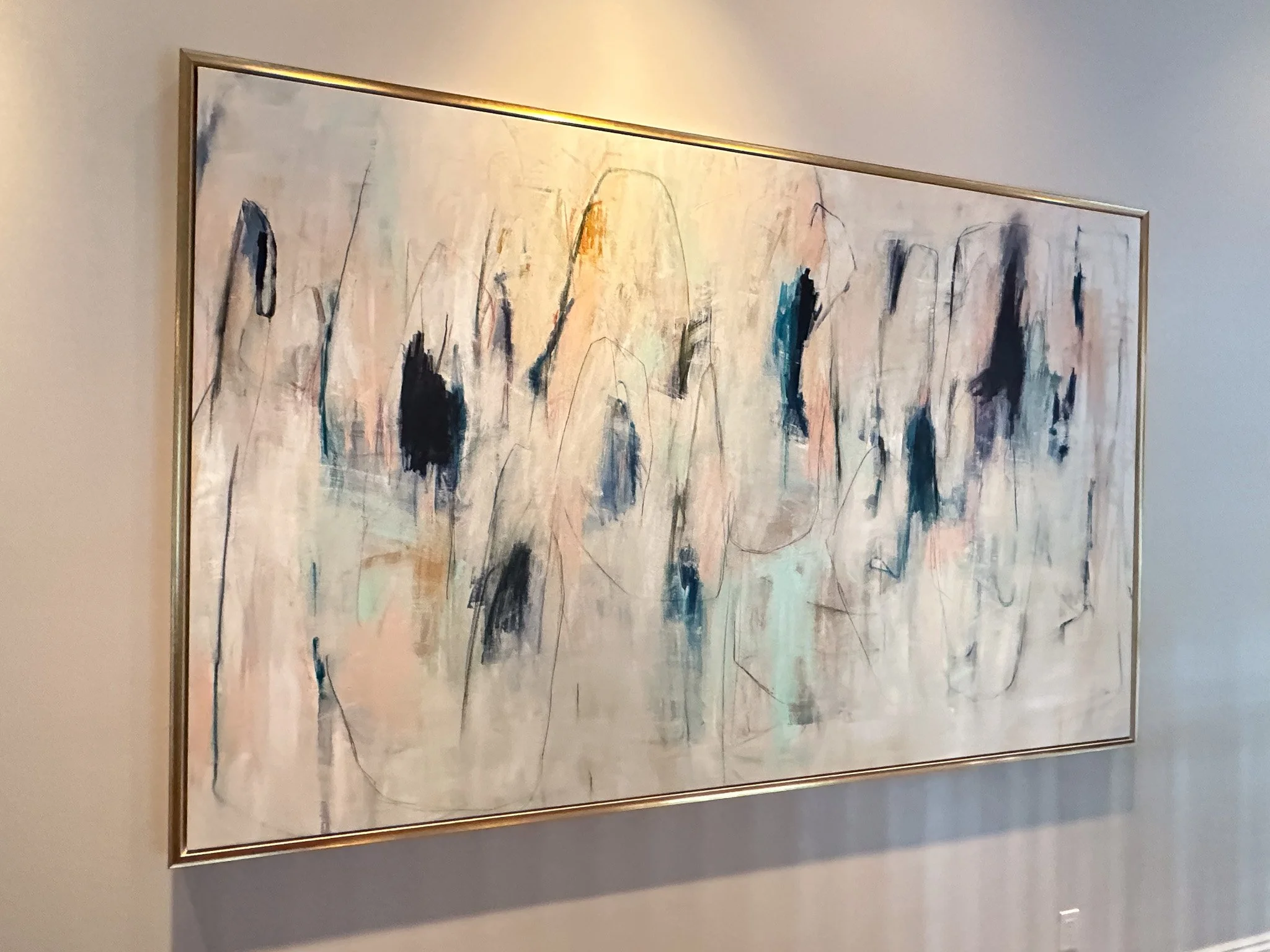 A large abstract painting with soft pastel colors and black and blue streaks, framed in a metallic frame, hanging on a wall.