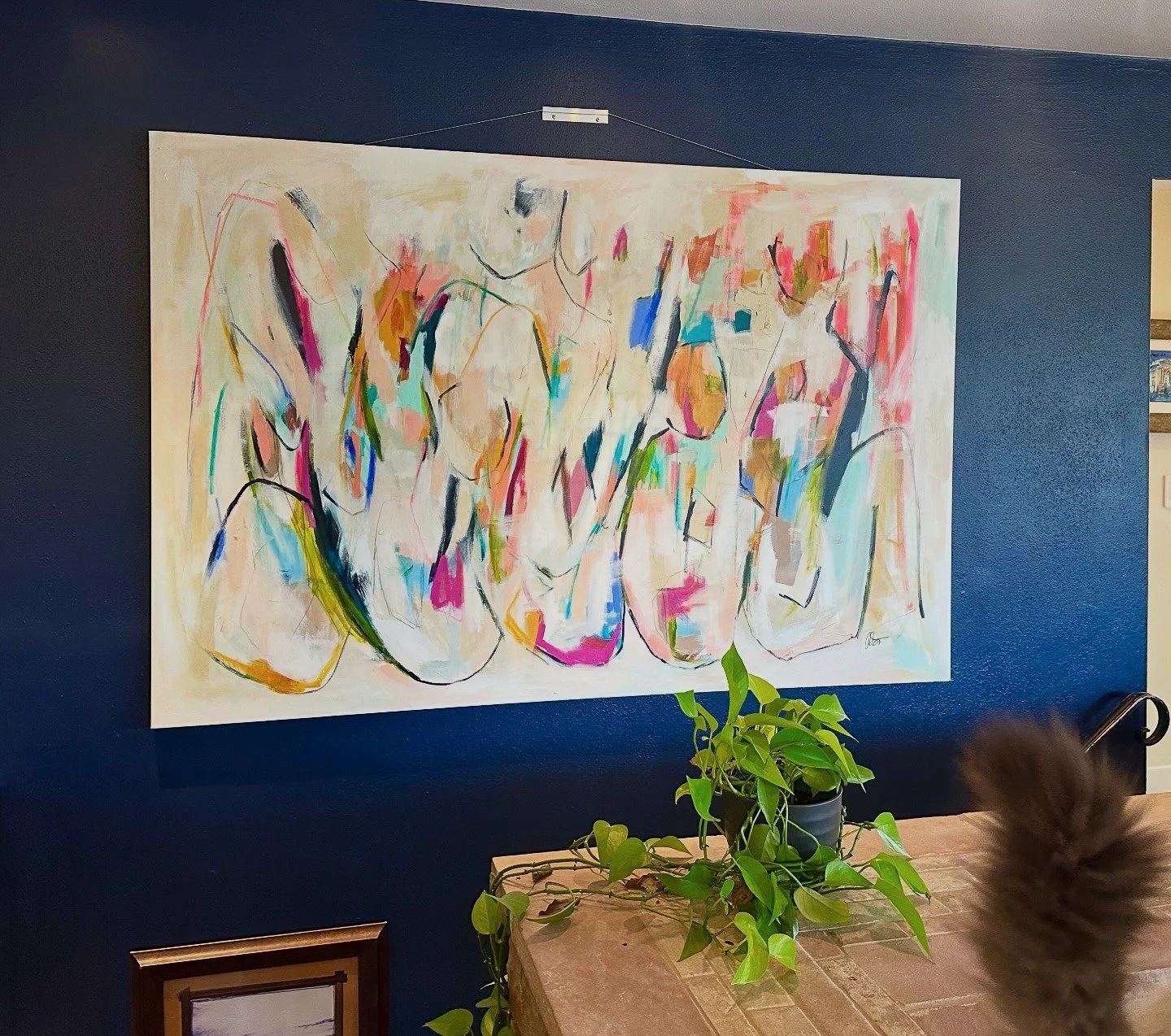 Abstract painting with colorful, flowing lines and shapes displayed on a dark blue wall. There is a wooden table in front with a potted green plant, and part of a person's head is visible at the bottom right.