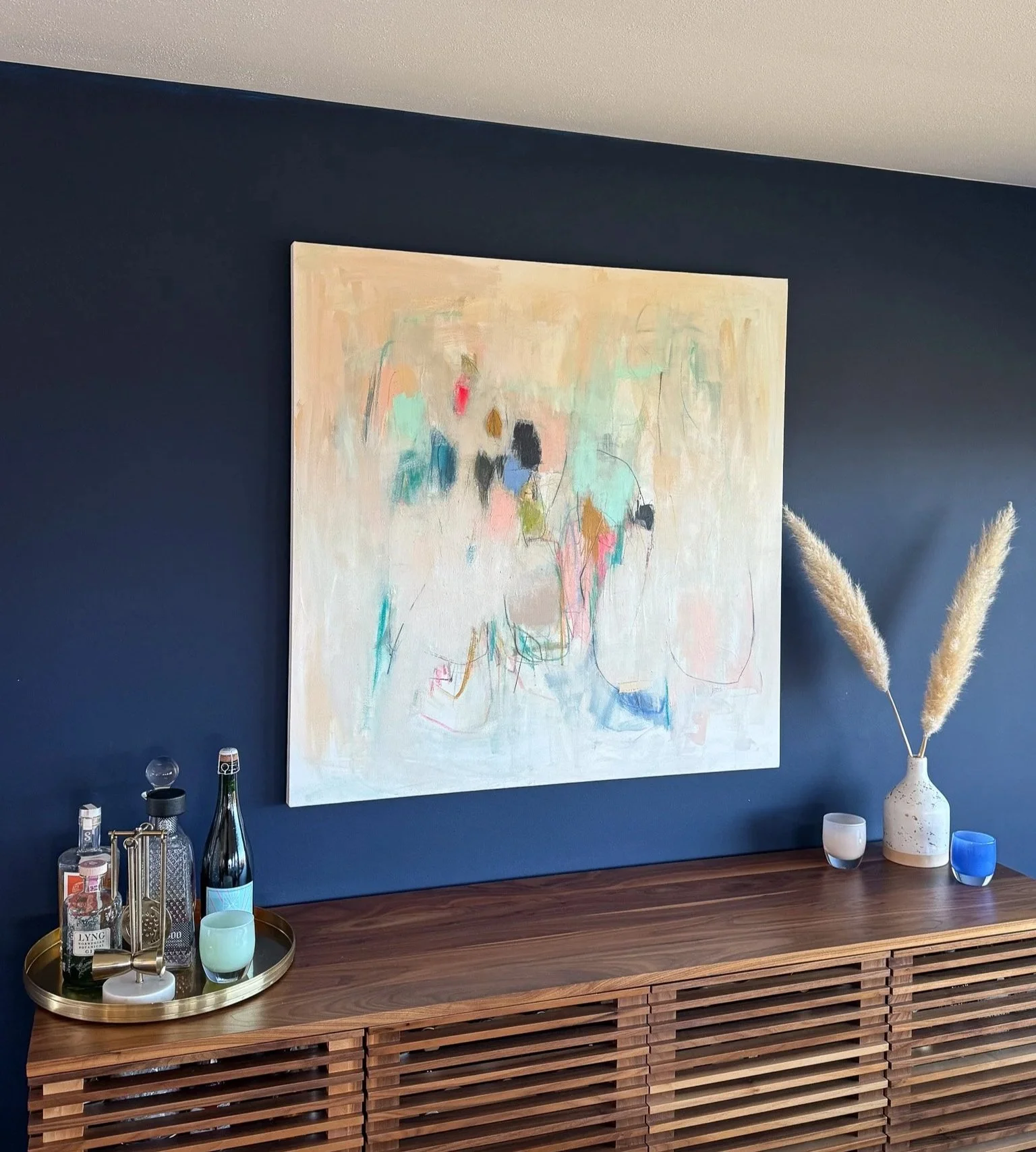 A modern wooden sideboard against a dark blue wall with a large abstract painting hanging above it. The sideboard holds a gold tray with various bottles and glasses, and has a beige vase with dried pampas grass on the right. There are small candles and decorative items on the sideboard.