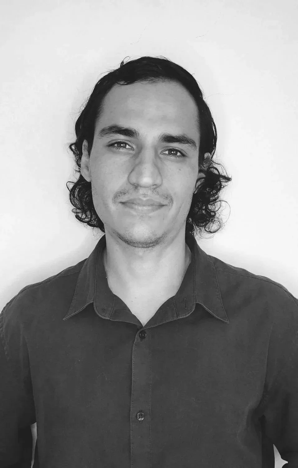 Retrato en blanco y negro de un hombre con cabello oscuro y rizado, sonriendo ligeramente, usando una camisa de botones oscura.