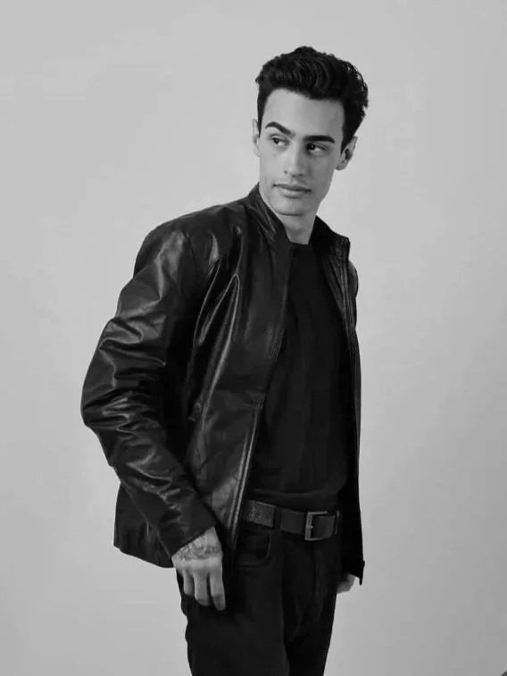 Joven con cabello oscuro y despeinado, lleva una chaqueta de cuero y ropa negra, mirando hacia la izquierda en un fondo gris.