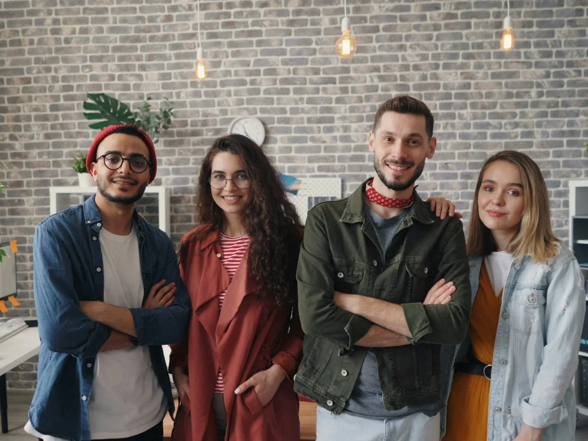 Cuatro jóvenes adultos sonparados y sonriendo en un ambiente de oficina moderna con paredes de ladrillo expuesto y lámparas colgantes.