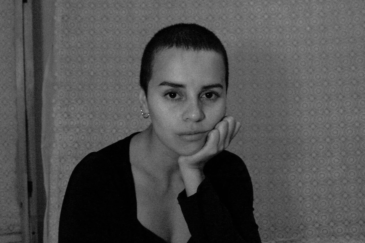 Retrato en blanco y negro de una mujer joven con cabello corto y cejas delgadas, posa con la cabeza apoyada en la mano, viendo hacia la cámara, en un fondo con patrón de círculos.