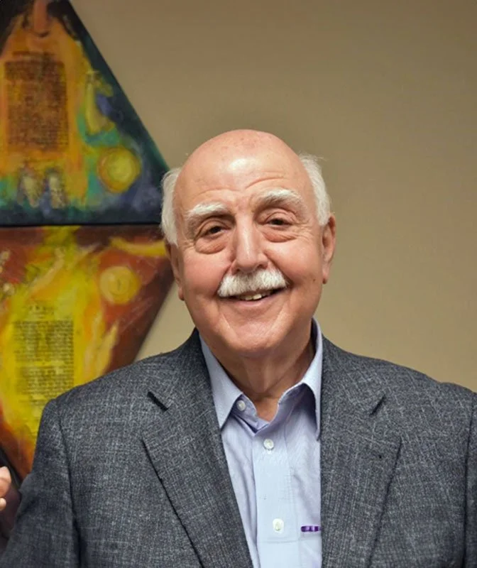 Howard Epstein- founder, dressed in a gray blazer and light blue shirt, standing in front of colorful abstract art on a beige wall.