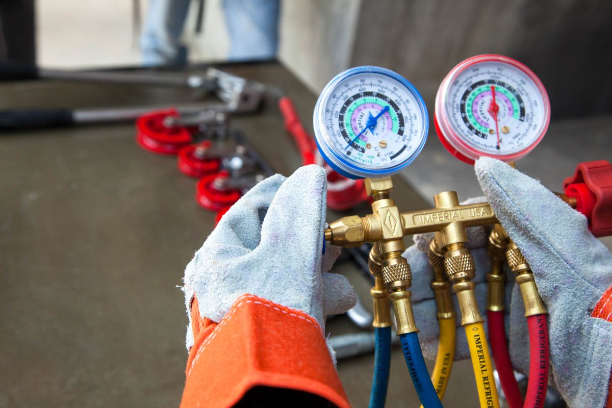 Close-up of a technician's gloved hands holding a manifold gauge set connected to refrigerant lines, with gauges displaying pressure readings, on a workbench in an HVAC lab.