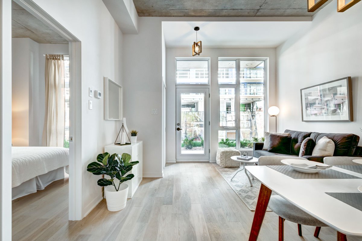 Bright modern living room with large windows, a dark sofa with pillows, a white armchair, a round lamp on a stand, a coffee table, and a patterned rug.