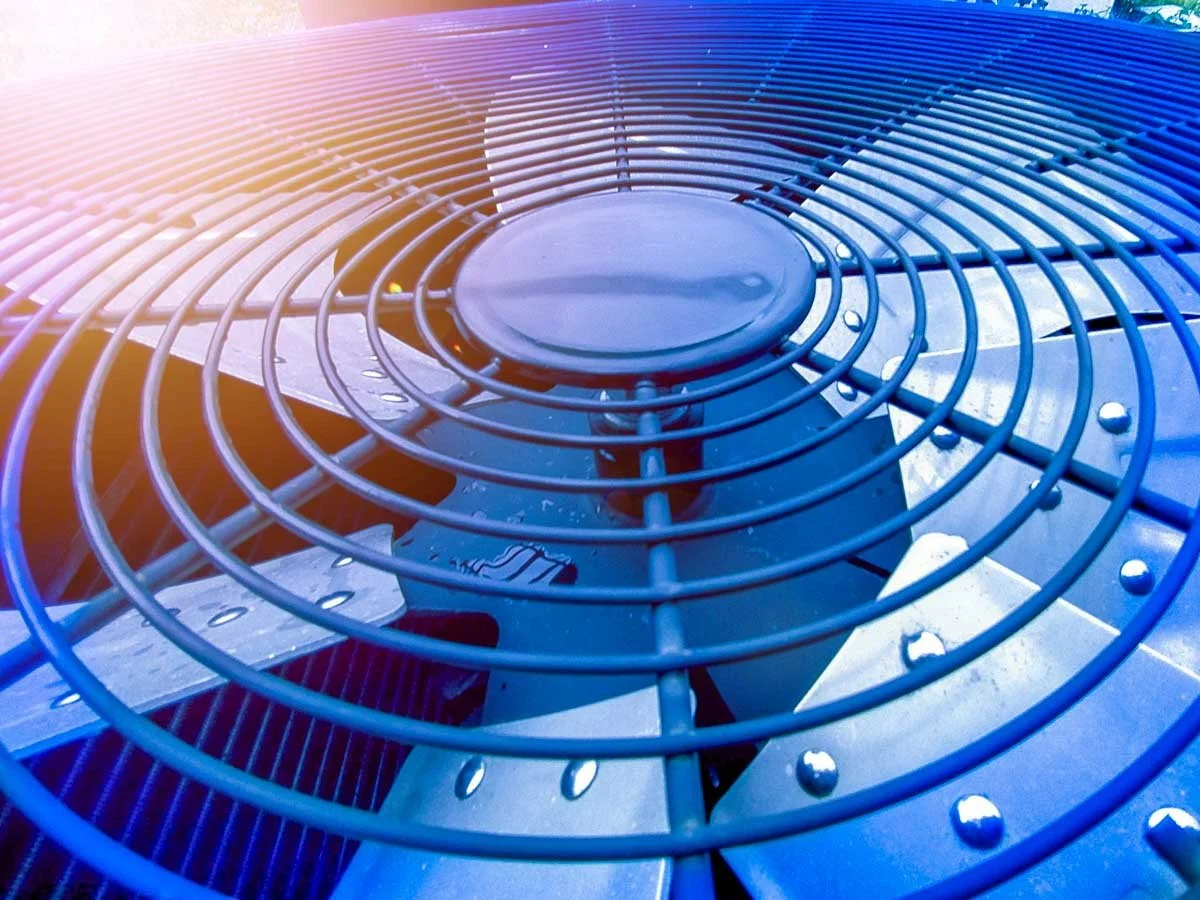 Close-up of an industrial air conditioning or HVAC unit's fan grill with metal blades and protective metal cage, with a gradient of warm pink and cool blue lighting.