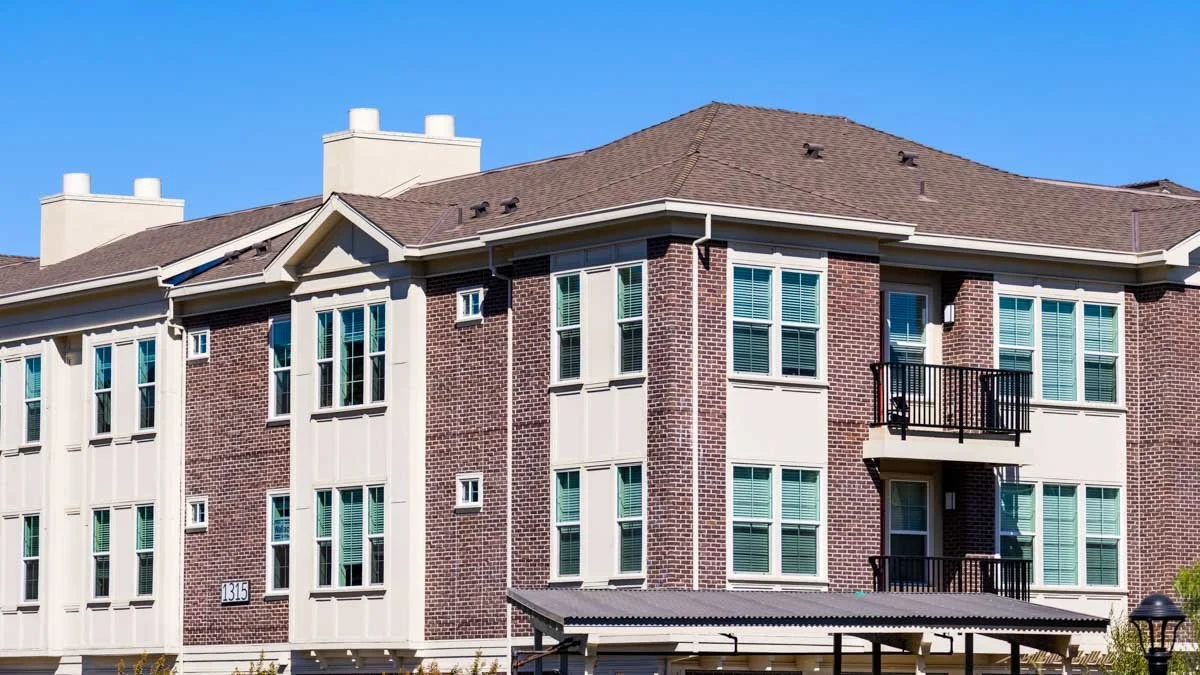 A multi-story residential building with brick and beige siding, multiple windows, and small balconies, under a clear blue sky.