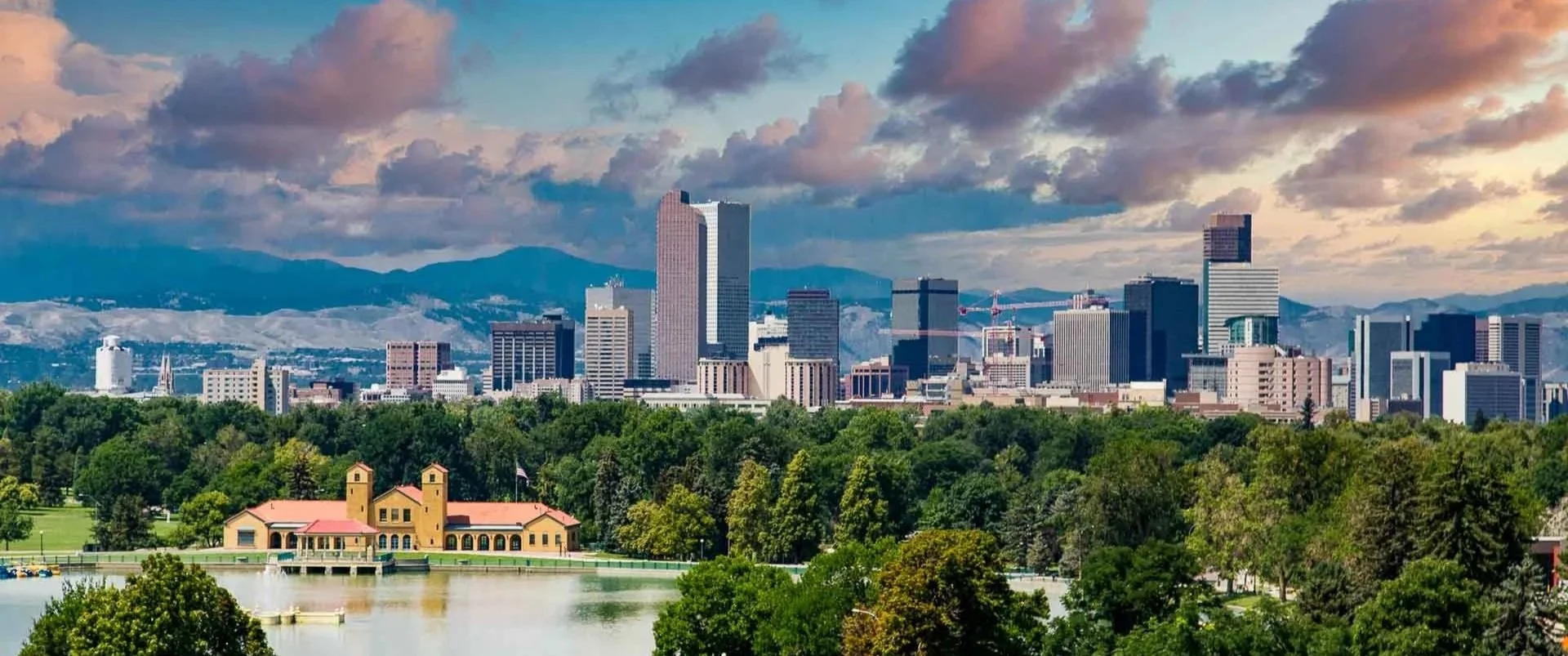 City skyline with tall buildings and skyscrapers, a park with trees and a pond in the foreground, and mountains in the background under a partly cloudy sky.