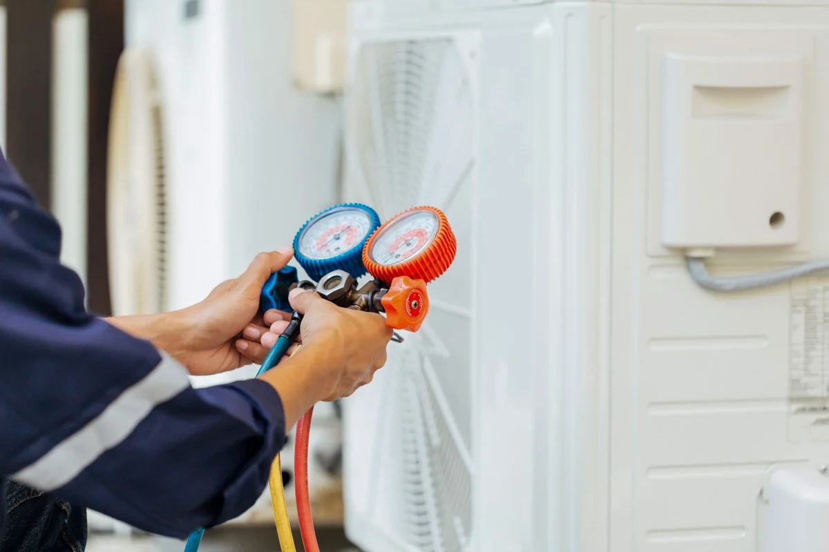 Person holding refrigeration manifold gauge set with two gauges and color-coded hoses next to an appliance.