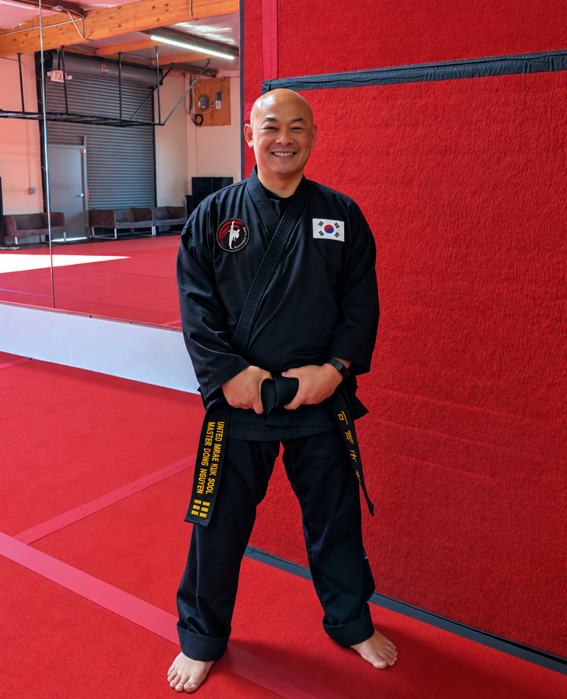 Master Dong Nguyen in uniform standing with his hands clasped in front of him inside a martial arts studio with red mats and a red wall.