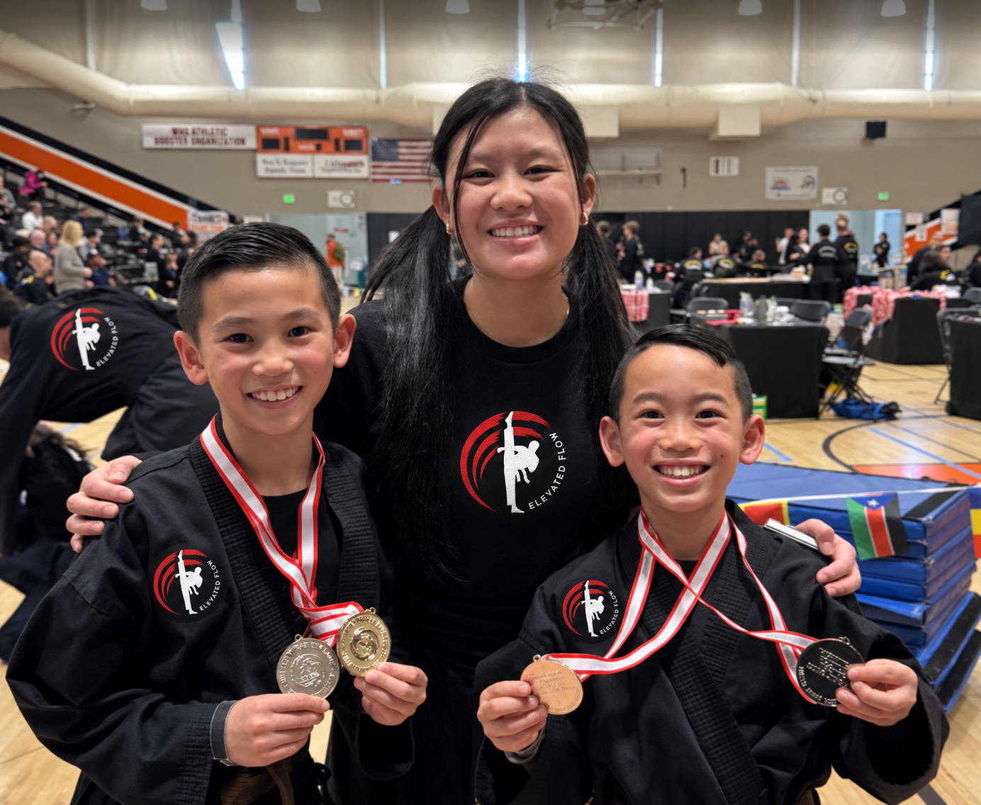 A woman and two boys smiling in a gymnasium, all wearing black martial arts uniforms with a logo that says 'Elevated Flow,' and medals around their necks, celebrating a martial arts competition.