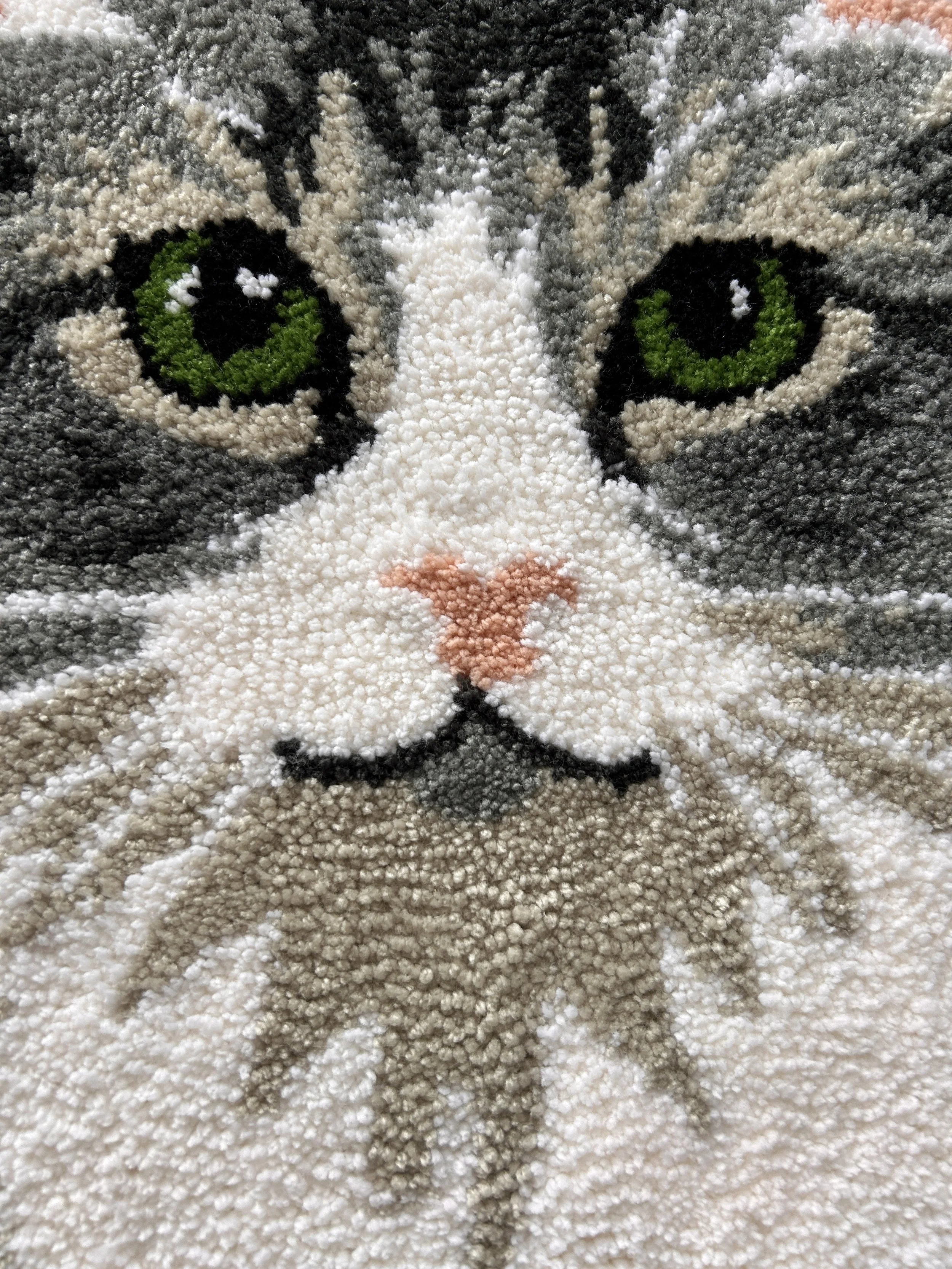 Close-up of a textured, plush rug with a detailed image of a kitten's face, showing bright green eyes, black and white fur, pink nose, and whiskers.
