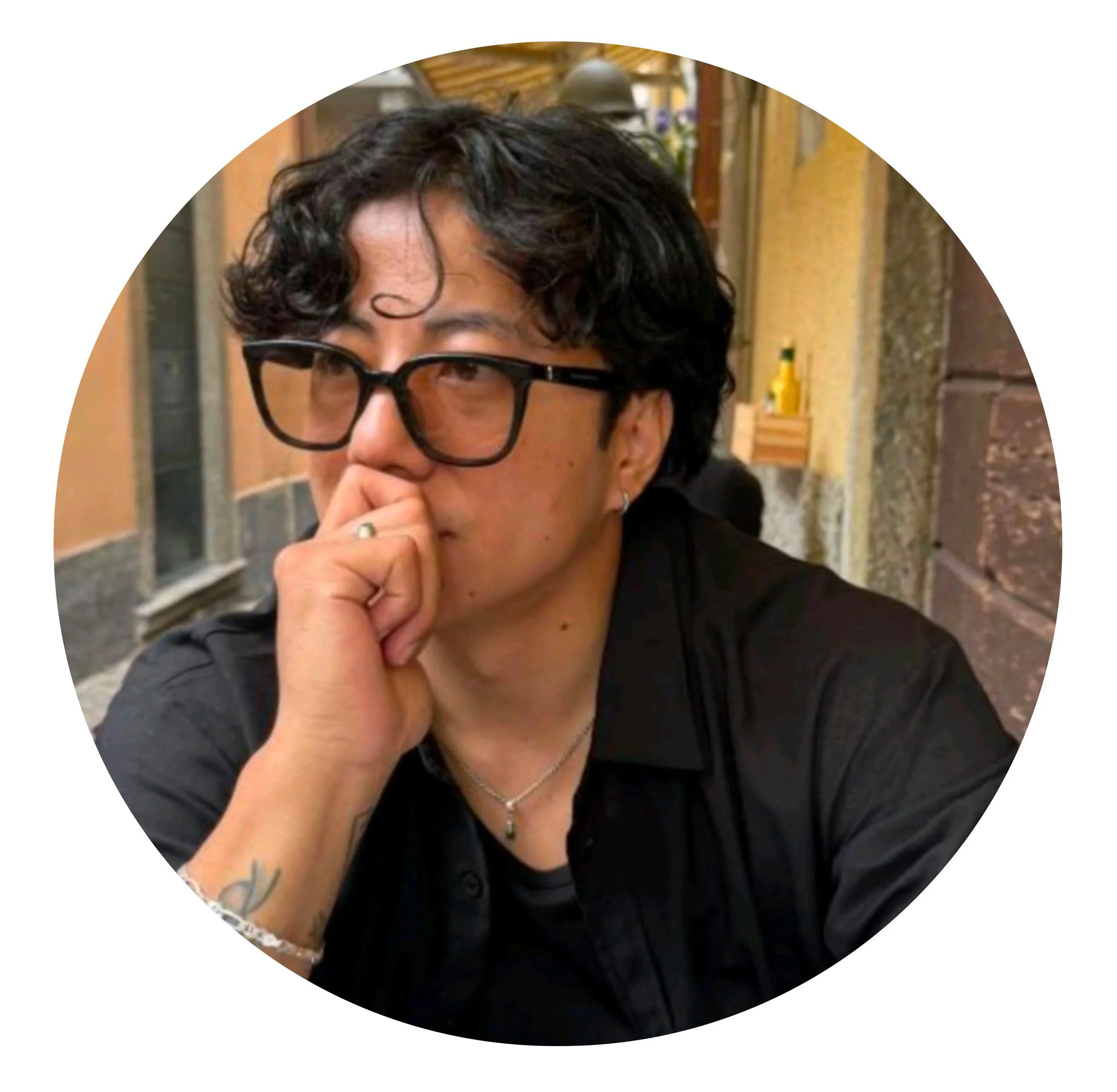 A person with dark, curly hair, black glasses, and light skin resting their chin on their hand at an indoor location.