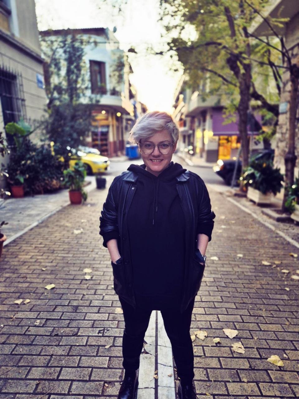 A woman with short blond hair, glasses, and a black outfit standing on a cobblestone street with trees and buildings in the background during daytime.