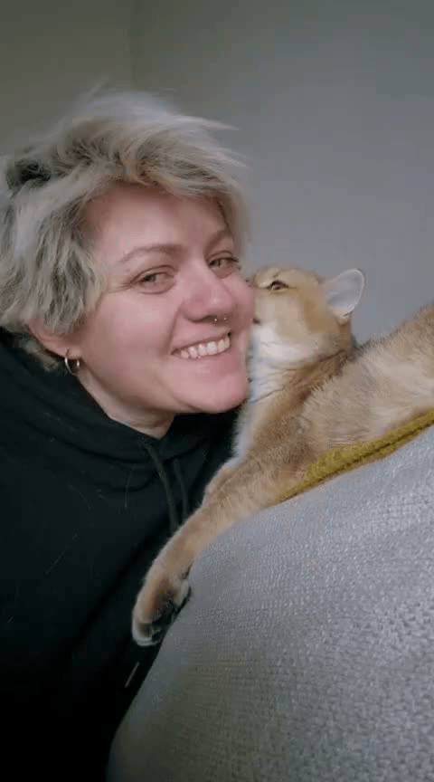 A woman with short blonde hair and a nose ring smiling as she kisses a ginger cat.