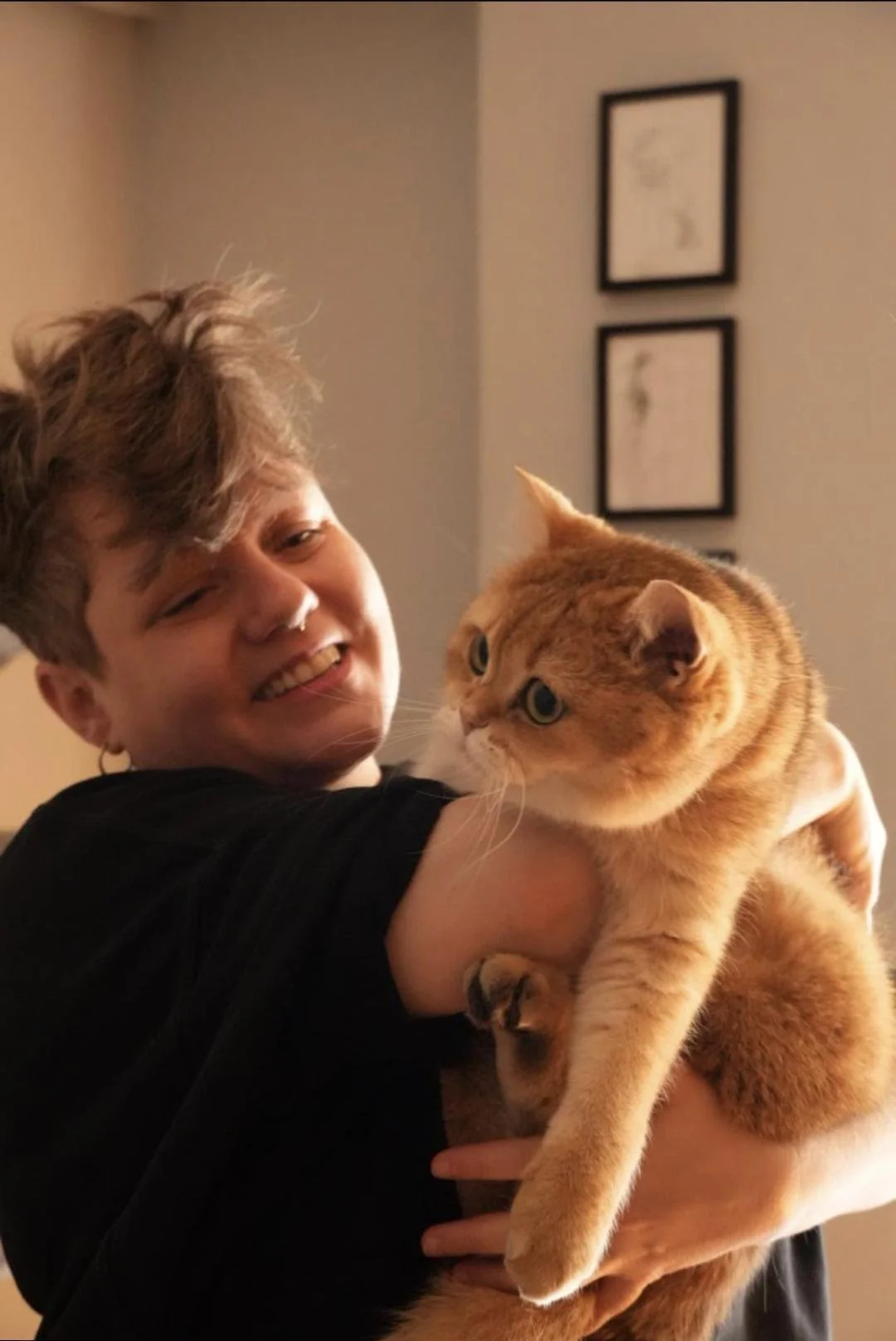 A young person with short curly hair smiling and holding a fluffy orange tabby cat with blue eyes indoors, with framed pictures on the wall in the background.