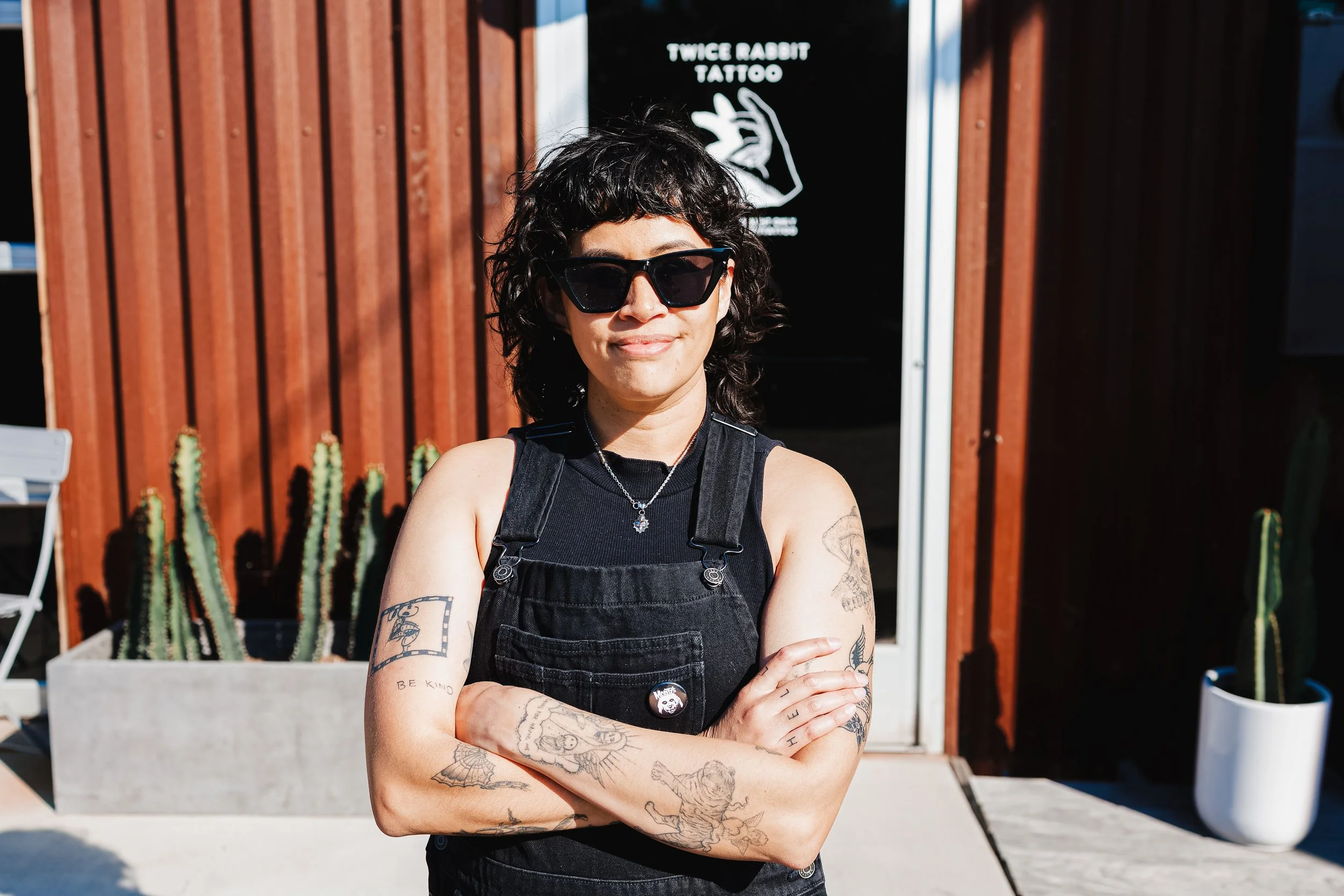 A woman with curly black hair, wearing sunglasses and a black sleeveless top, standing outdoors with arms crossed. She has tattoos on her arms and is smiling slightly. In the background, there is a sign for a tattoo shop called 'Twice Rabbit Tattoo' and some cacti.
