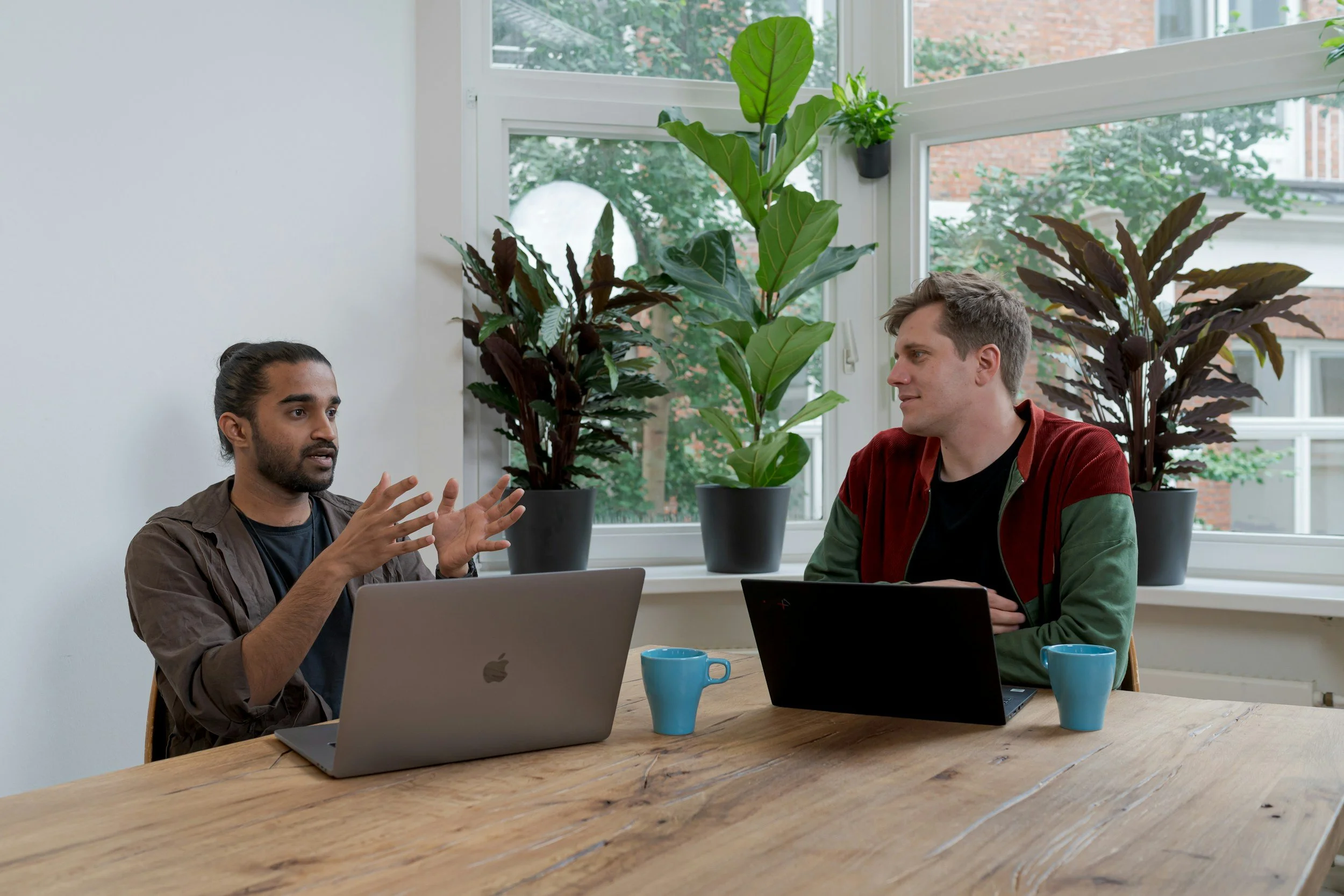 Two men engaged in a conversation at a wooden table in a bright room with large windows. Potted plants and greenery are visible outside the window, and two blue mugs and two laptops are on the table.