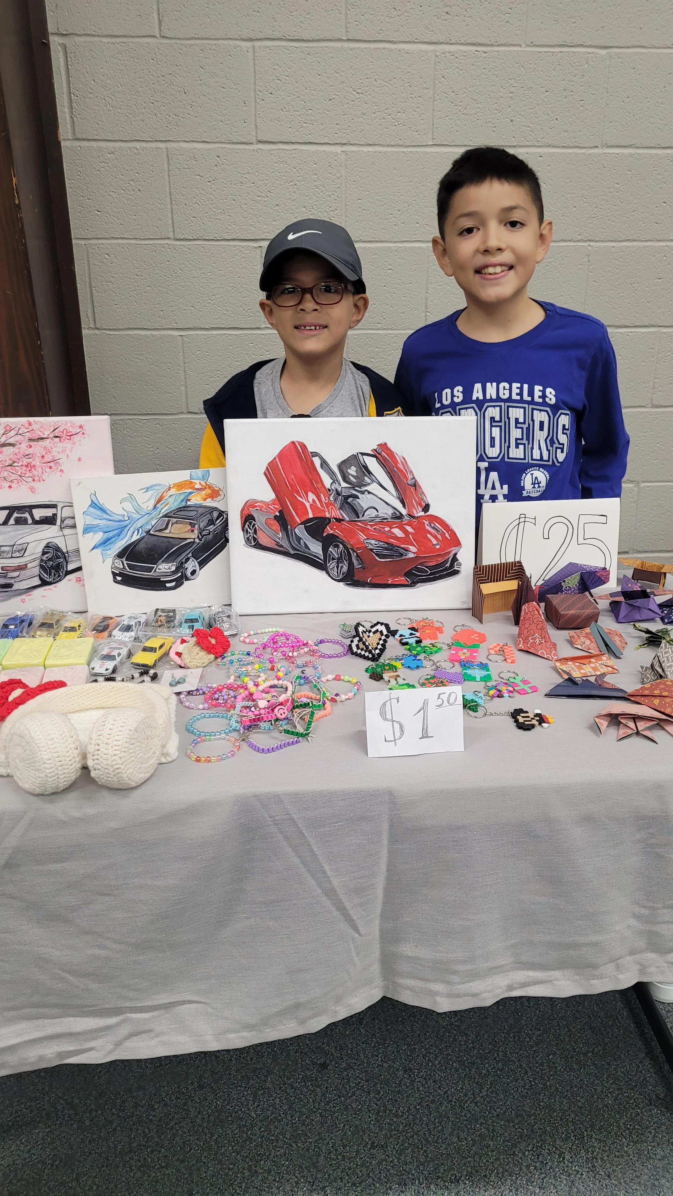 A table with jewelry, paper crafts, and artwork displayed for sale at a craft fair, with two boys behind it, one wearing glasses and a cap, and the other wearing a blue shirt.