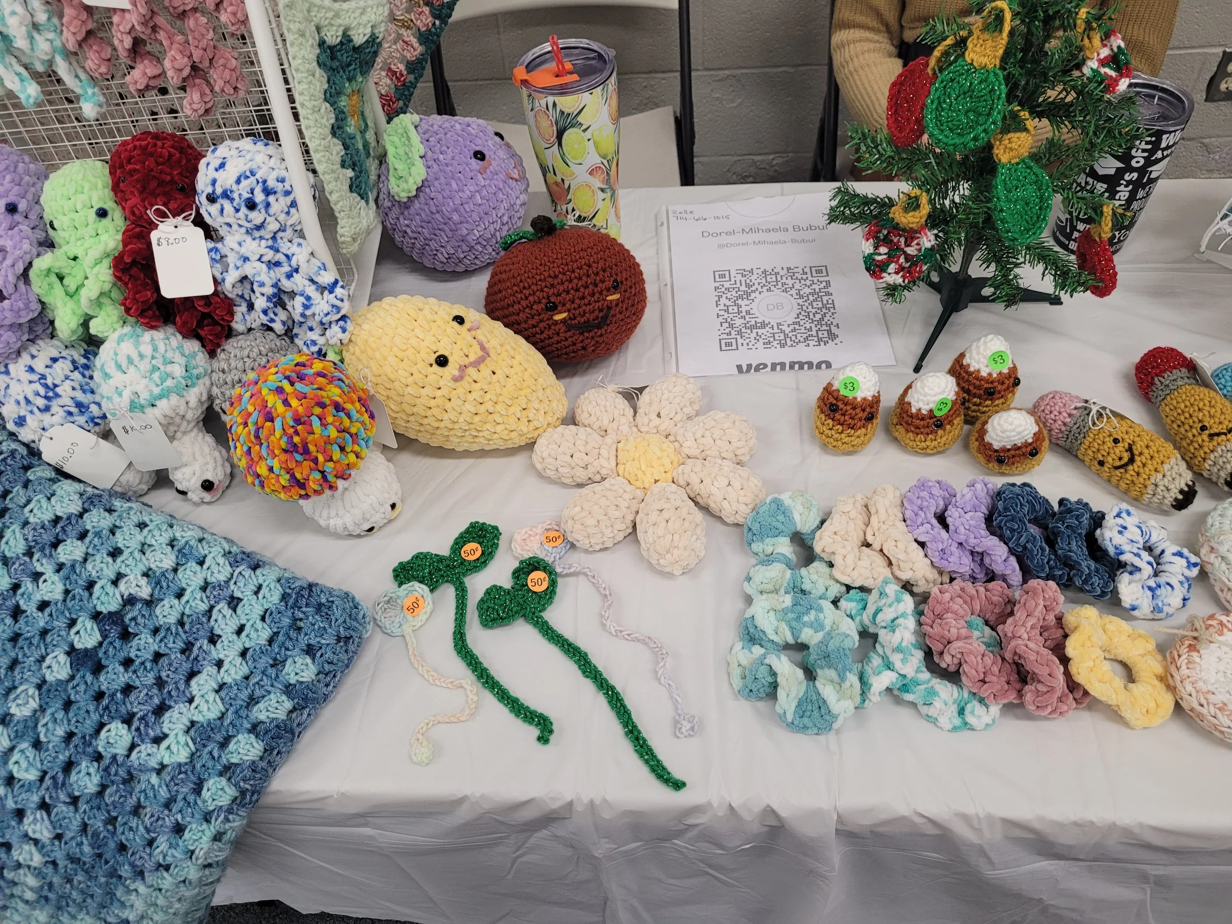 A table displaying various handmade crochet items, including stuffed animals, pumpkins, and decorative ornaments, with price tags. There is a small artificial Christmas tree decorated with crochet ornaments. A QR code and sign with contact information are also on the table.