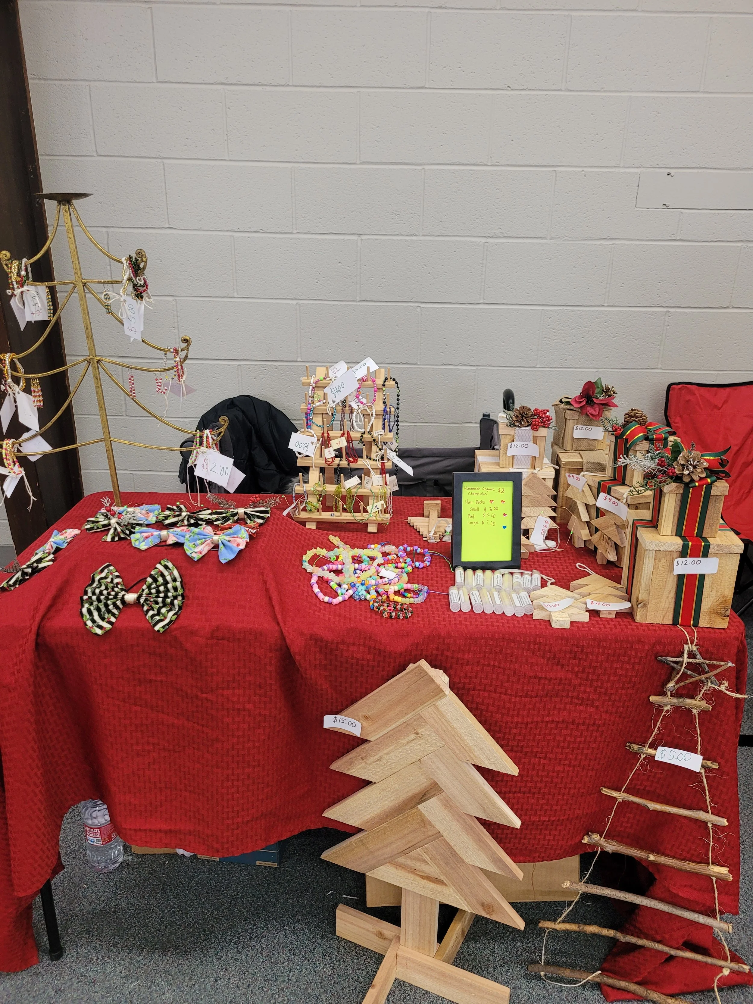 A table covered with a red cloth displaying handmade jewelry, including bows and beaded necklaces, and wooden crafted items on the right side, some with price tags, in a market setting against a beige cinder block wall.