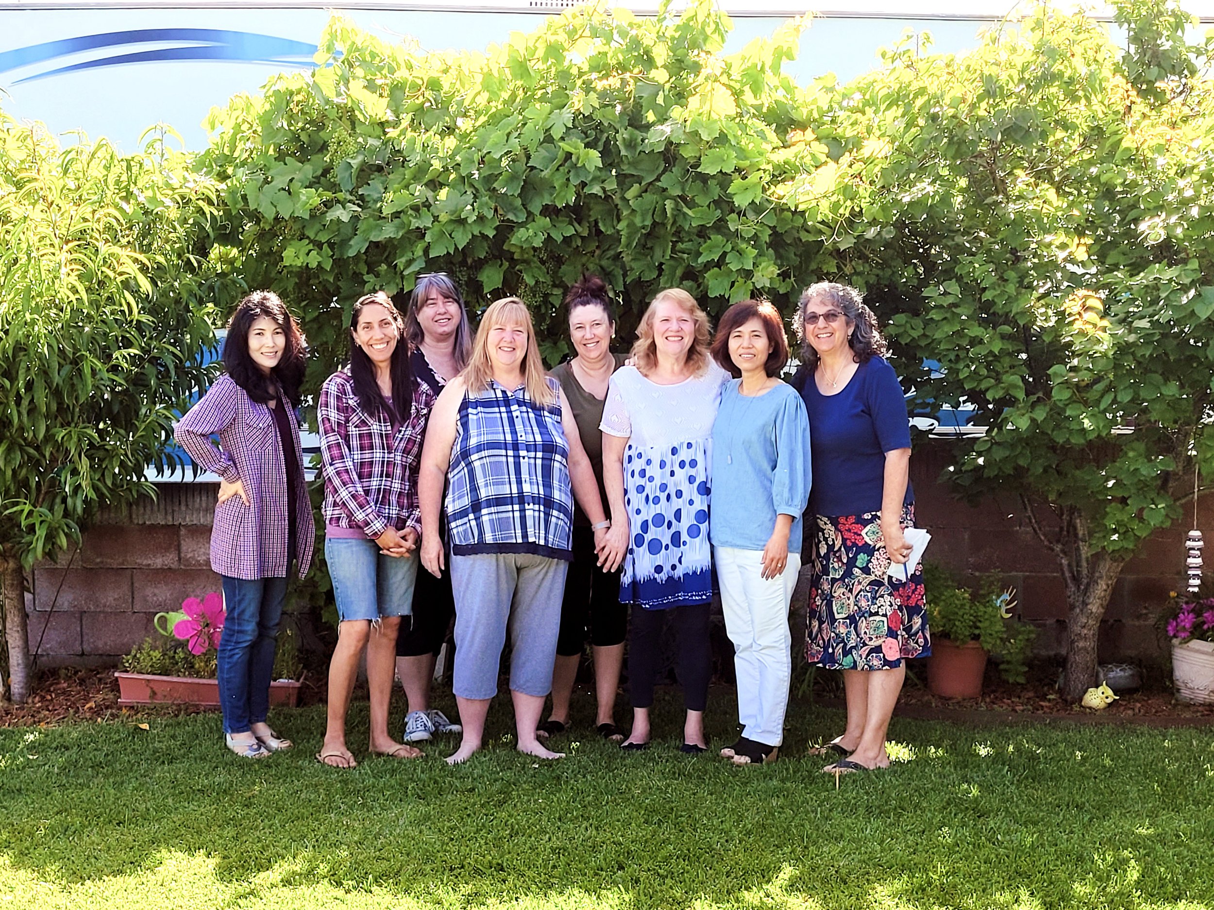 Group of nine women standing together outside in a garden, smiling. They are surrounded by green trees, potted plants, and flowers, with bright sunlight.
